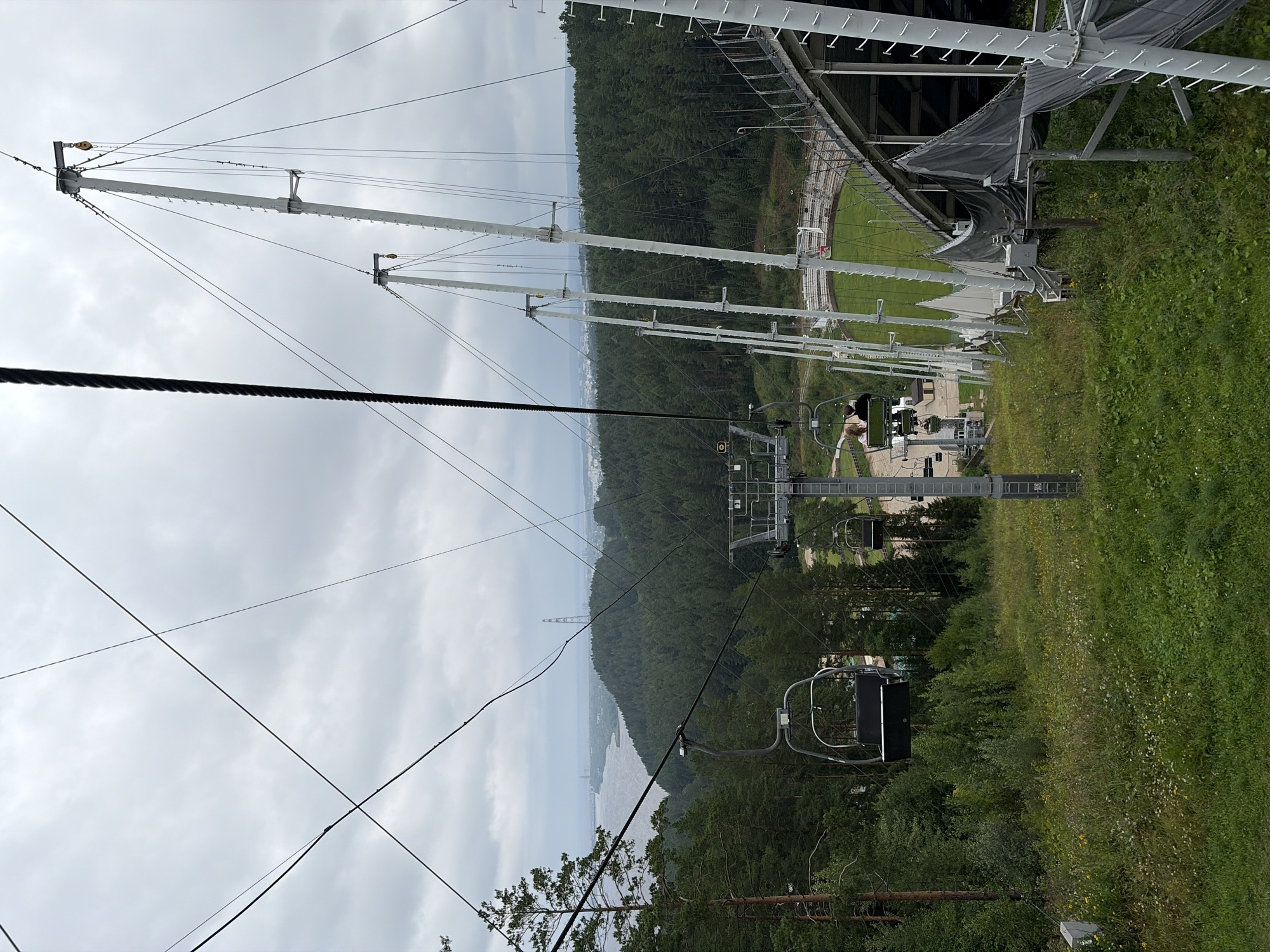 The image depicts a ski lift operating on a cloudy day. In the foreground, a grassy slope with scattered wildflowers leads down to a ski jump facility. The ski jump structure is large and white, with a ramp that curves downwards. Several white support poles for the ski lift are visible, extending upwards towards a thick cable that runs across the sky.
Two ski lift chairs are visible. The closest chair is empty and suspended mid-air. Further down the slope, another chair carries two individuals, a man and a woman, seated side-by-side.
In the background, a dense forest of green trees covers rolling hills. Beyond the forest, a wide river or bay stretches out, with a distant cityscape and a tall transmission tower visible on the horizon. The overall atmosphere is subdued due to the overcast sky. The location is identified as the ski jump facility in the city of Perm, Russia, specifically in the area of Прикамский.
