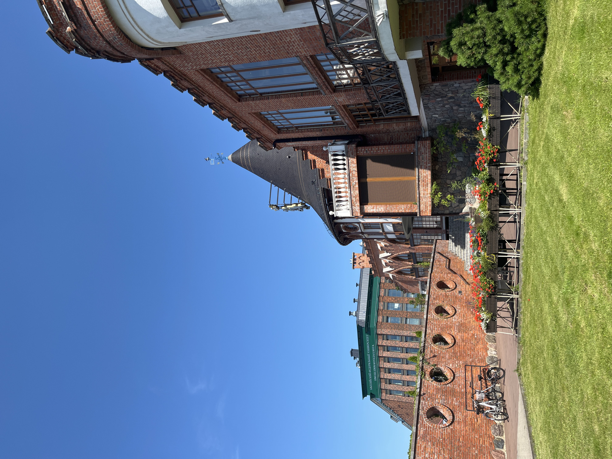The image captures a sunny day in Kaliningrad, Russia, showcasing a section of the Immanuel Kant Baltic Federal University's campus. The architecture is a blend of historical and modern styles. In the foreground, a lush green lawn leads to a red brick wall with circular openings. Two bicycles are parked near the wall. Beyond the wall, a multi-story building with a green roof and the university's name inscribed in white letters on it stands against a clear blue sky. Adjacent to this is a building with a distinctive conical roof, featuring a weather vane. The buildings are constructed primarily of red brick and stone, with large windows and balconies. Flower boxes filled with red and white flowers are arranged in front of a covered walkway. The overall scene suggests a well-maintained and aesthetically pleasing campus environment.