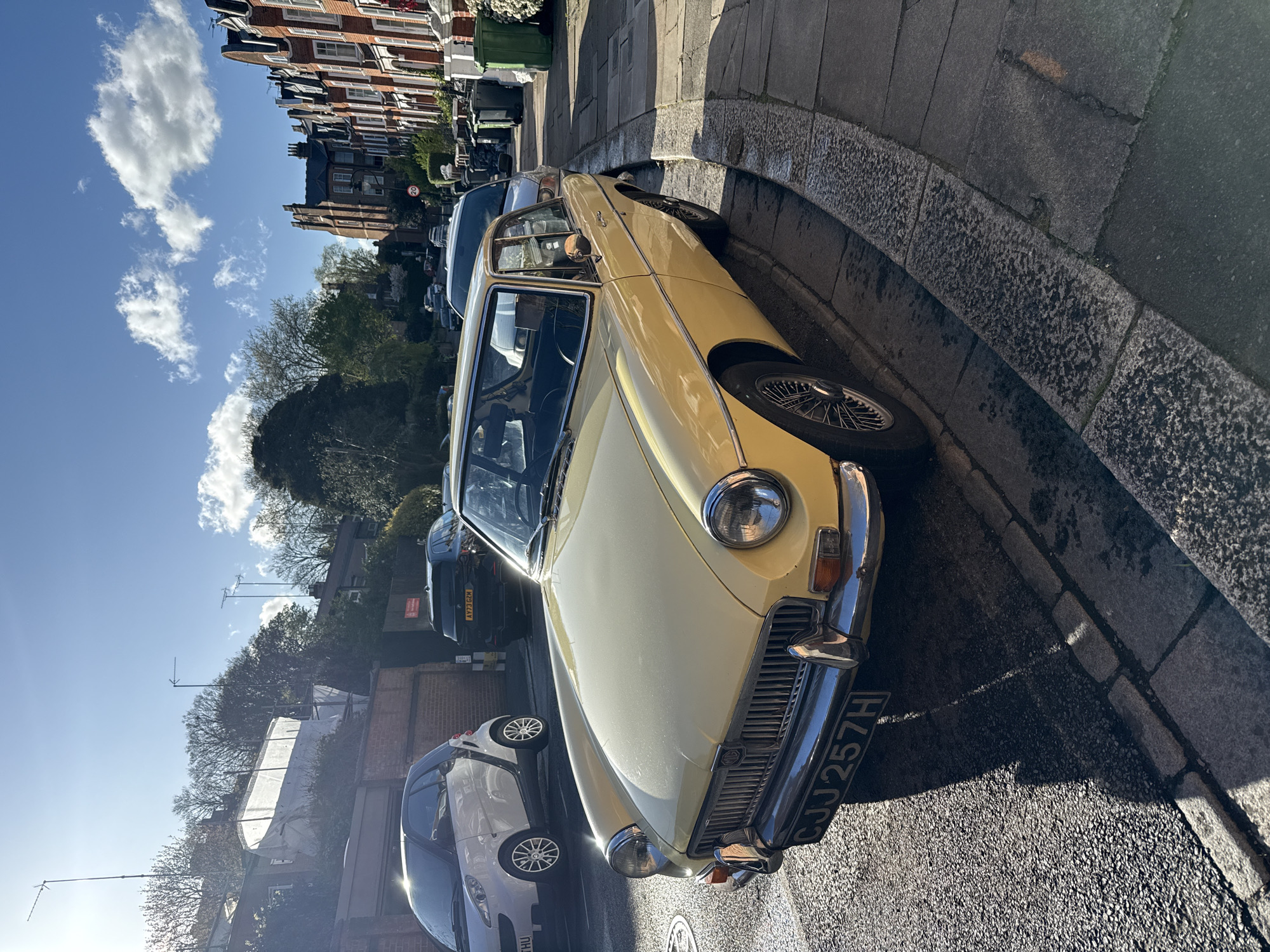 The image depicts a yellow MGB roadster parked on a residential street in London, United Kingdom. The car is positioned on the curb along a bend in the road. Other vehicles, including a silver Fiat 500 and a dark-colored SUV, are parked further down the street. The houses lining the street are constructed of brick. The sky is blue with scattered clouds. The scene is well-lit with sunlight casting shadows on the street and vehicles. A manhole cover is visible in the foreground near the front of the MGB. The license plate of the MGB reads CJJ 257H.