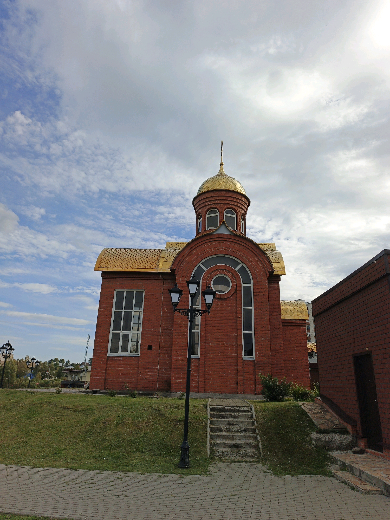 The image shows the Church of St. Sergius of Radonezh in Zlatoust, Russia. It is a small, red brick church with a golden dome and roof. The church is situated on a grassy hill with a paved walkway in the foreground. There are several streetlights around the church. The sky is cloudy.