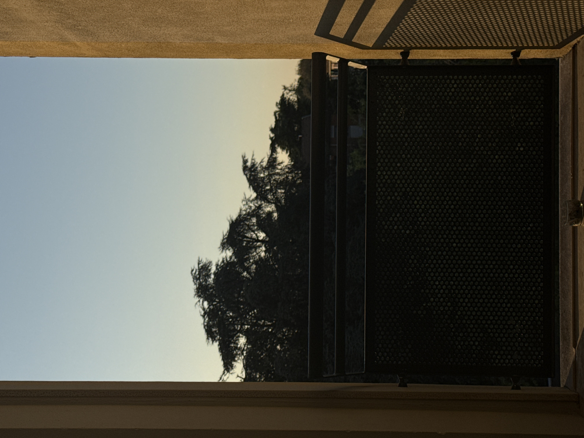 The image captures an outdoor scene, likely rotated 90 degrees clockwise from its original orientation, presenting a view looking upwards and outwards from beneath a building's structure. The upper half of the frame is filled with a clear sky, displaying a gentle gradient from pale blue at the top to a lighter, slightly warmer, possibly yellowish tone towards the horizon. This soft lighting suggests either an early morning sunrise or a late afternoon sunset.
Silhouetted against the bright sky on the right side are the dark, intricate forms of dense trees, their leafy branches standing out. The lower portion of the image reveals architectural elements. An off-white or light brown textured surface, possibly the underside of a balcony or overhang, spans the bottom. Sharp, geometric shadows, cast from a grid-like pattern (likely a railing or pergola above), are distinctly visible on this surface, indicating direct sunlight.
On the right, a dark, perforated metal panel with numerous small circular holes forms part of a railing or decorative screen. Behind this panel, several vertical slats reflect light. The overall scene is framed by elements of the building, suggesting a view from a balcony, terrace, or window in an urban or suburban environment. There is no visible activity, people, or text. The setting is Milan, Italy, and the atmosphere appears calm under a clear sky.