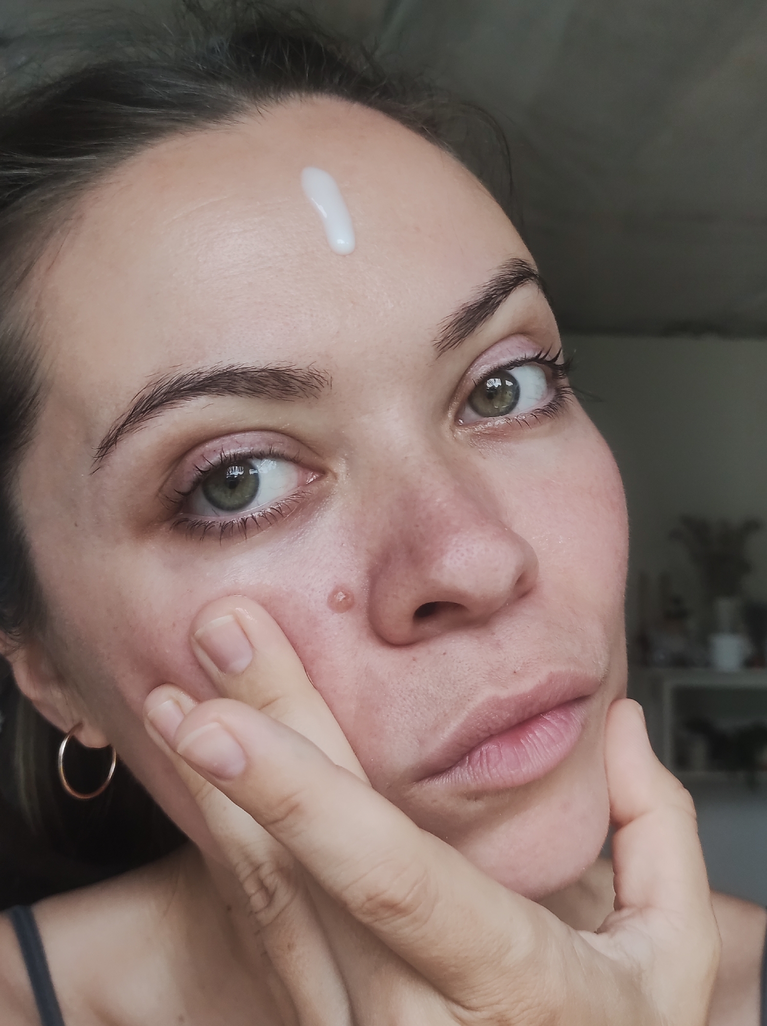 A close-up, head-on shot depicts a female with fair to medium skin tone, dark hair, dark eyebrows, and green/hazel eyes, looking directly at the camera. A single dollop of white, viscous product is positioned on her forehead. Her left hand's index finger is gently touching her right cheek below the eye, near a small, raised, flesh-toned mole. Her right hand is positioned against her left cheek and jawline. She wears a small, thin, gold-colored hoop earring in her left ear and a dark tank top. The background is out of focus, appearing dark on the left and showing indistinct shapes resembling furniture or shelves in a lighter tone on the right, indicating an indoor setting. The location in the city of Фоки, Russia, is not discernible from the image.