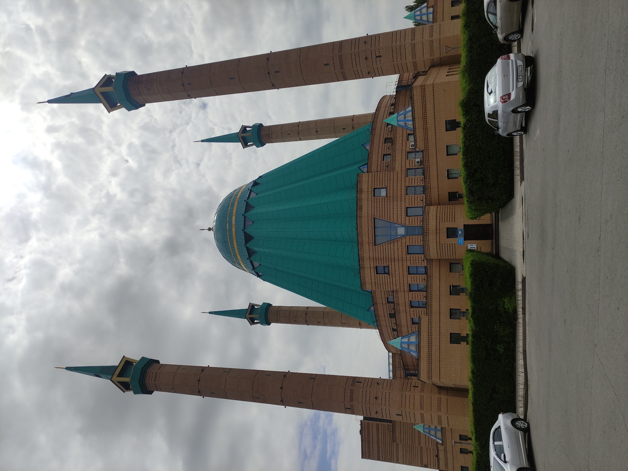 The image shows the Mashkhur Jusup Central Mosque in Pavlodar, Kazakhstan. The building is constructed of brown brick with a large, green, conical dome and four tall minarets topped with green spires. Two silver cars are parked along the side of the building on a paved road. A green hedge runs along the side of the mosque. The sky is overcast with grey clouds.