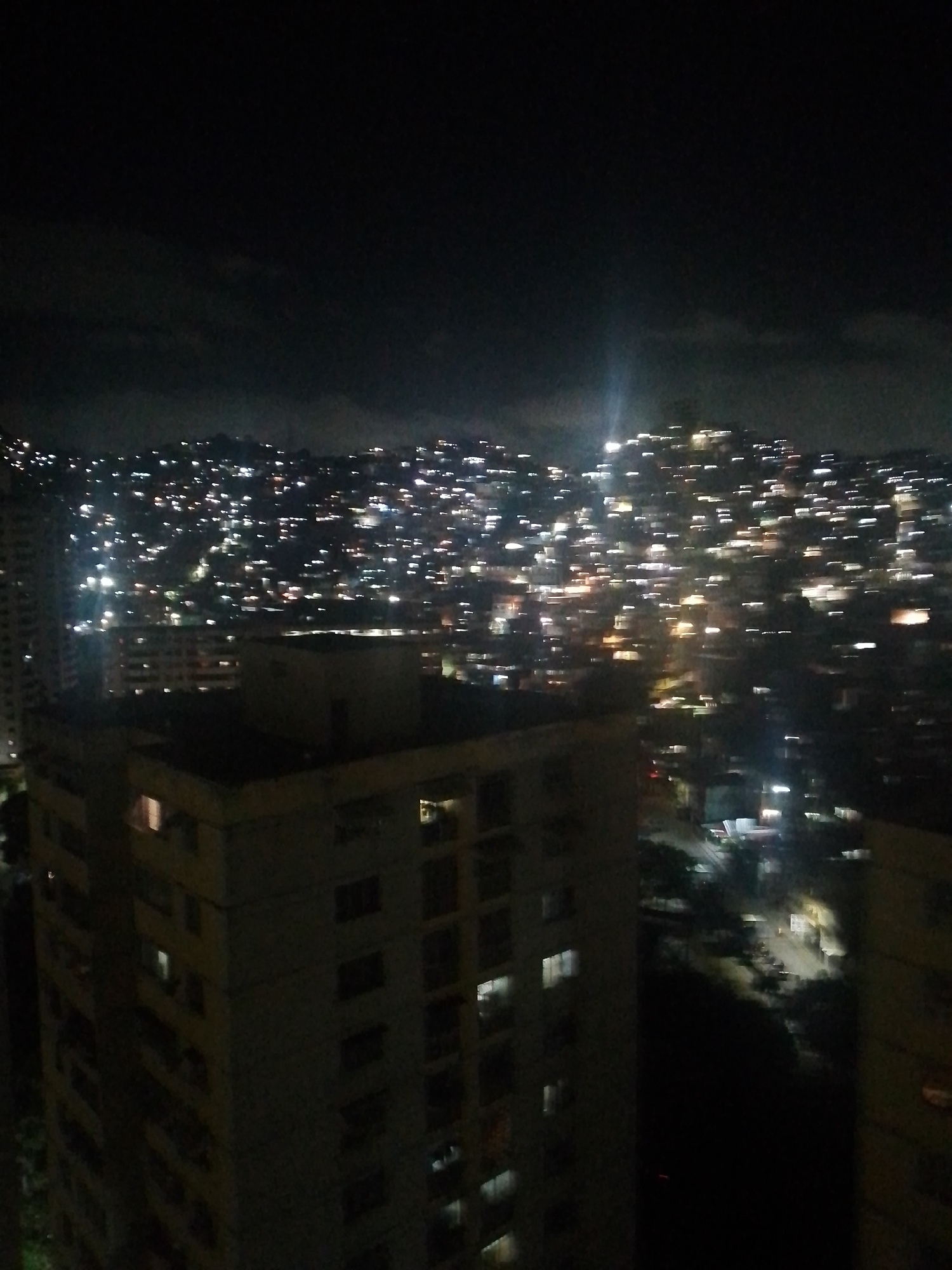 The image depicts a nighttime view of a densely populated hillside in Caracas, Venezuela, illuminated by countless lights from houses and buildings. The foreground features several multi-story apartment buildings, some with illuminated windows suggesting occupation. The hillside behind is a sprawling panorama of urban development, with lights cascading down the slopes, indicating a significant population density. The sky is dark, with some clouds visible. The overall impression is one of a vibrant, active city at night, characterized by its hilly terrain and extensive residential areas.