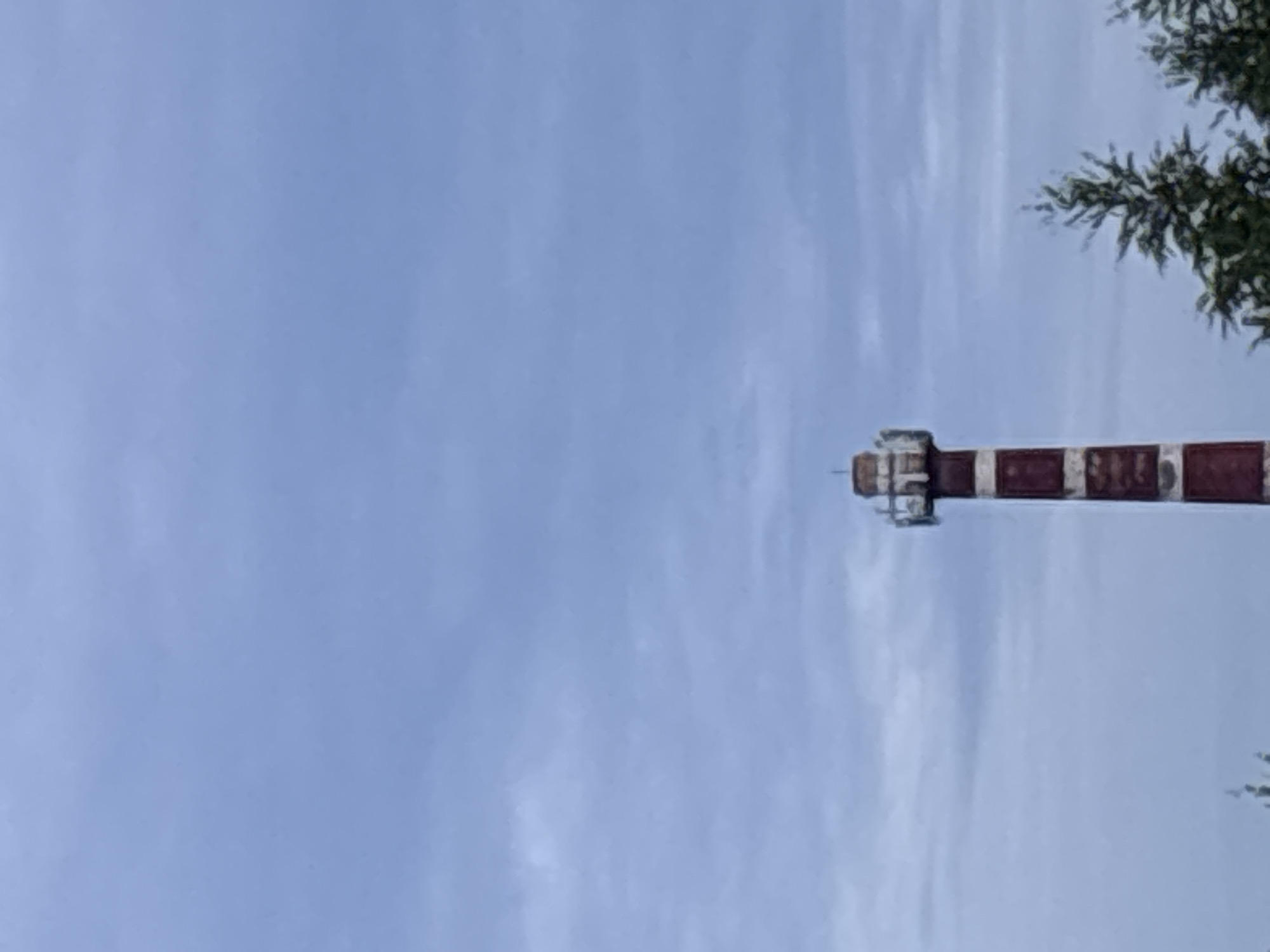 The image shows a red and white striped lighthouse against a cloudy sky. The lighthouse is tall and slender, with a platform at the top. There are some trees visible in the foreground, to the right of the lighthouse. The overall scene is overcast and muted in color. The lighthouse appears to be located in Zvenigorod, Russia, as indicated by the prompt.