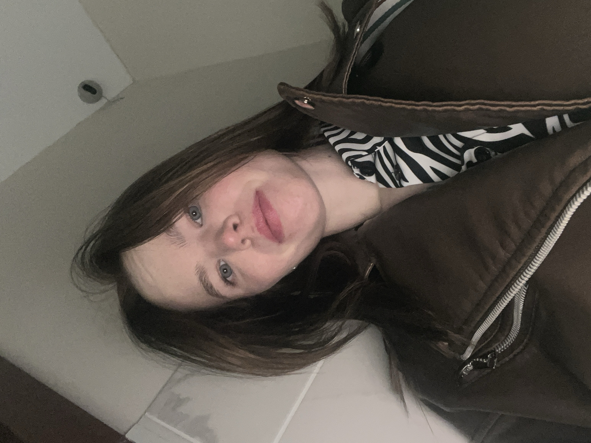 The media file is an angled, close-up indoor selfie of a young individual, taken in Chaikovsky, Russia. The subject, likely female, has long, straight dark brown hair, blue eyes, and a fair complexion. She is looking directly into the camera with a soft, slightly smiling expression and light pink lips.
She is dressed in a dark brown, possibly leather or faux leather, jacket with visible silver zipper hardware. Underneath the jacket, a black and white zebra-print top is visible around the neckline. The perspective is from a low angle, with the subject's head tilted, and her hair partially obscures the lower part of the frame.
The background consists of plain, light-colored walls and a ceiling. In the upper left corner of the image, a small, dark, circular object, resembling a smoke detector or a recessed light fixture, is mounted on the ceiling. The lighting appears soft and diffused, typical of indoor conditions. No specific activity beyond posing for the photo is evident, and the subject appears relaxed. There is no visible text in the image.