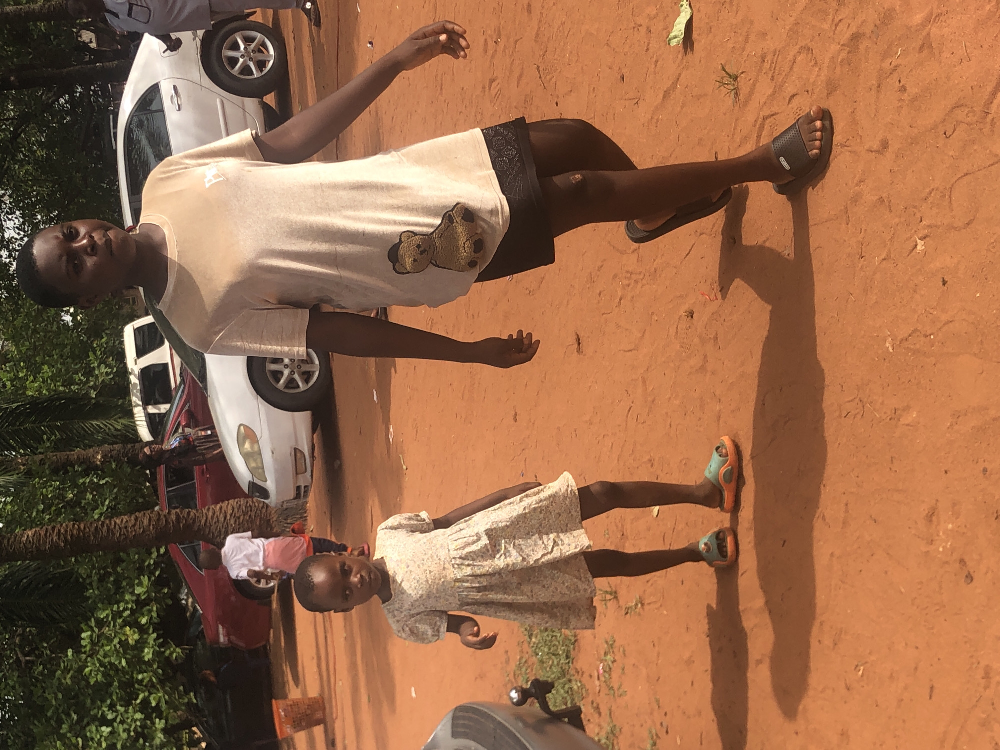 Two individuals are positioned on a dirt surface. The first individual, an adult, is wearing a light-colored t-shirt with a bear image, black shorts, and dark sandals. The second, a child, is wearing a light-colored floral dress and light-colored sandals. The background includes partial views of two cars, one white and one red, along with trees. The location is determined to be Nteje, Nigeria.