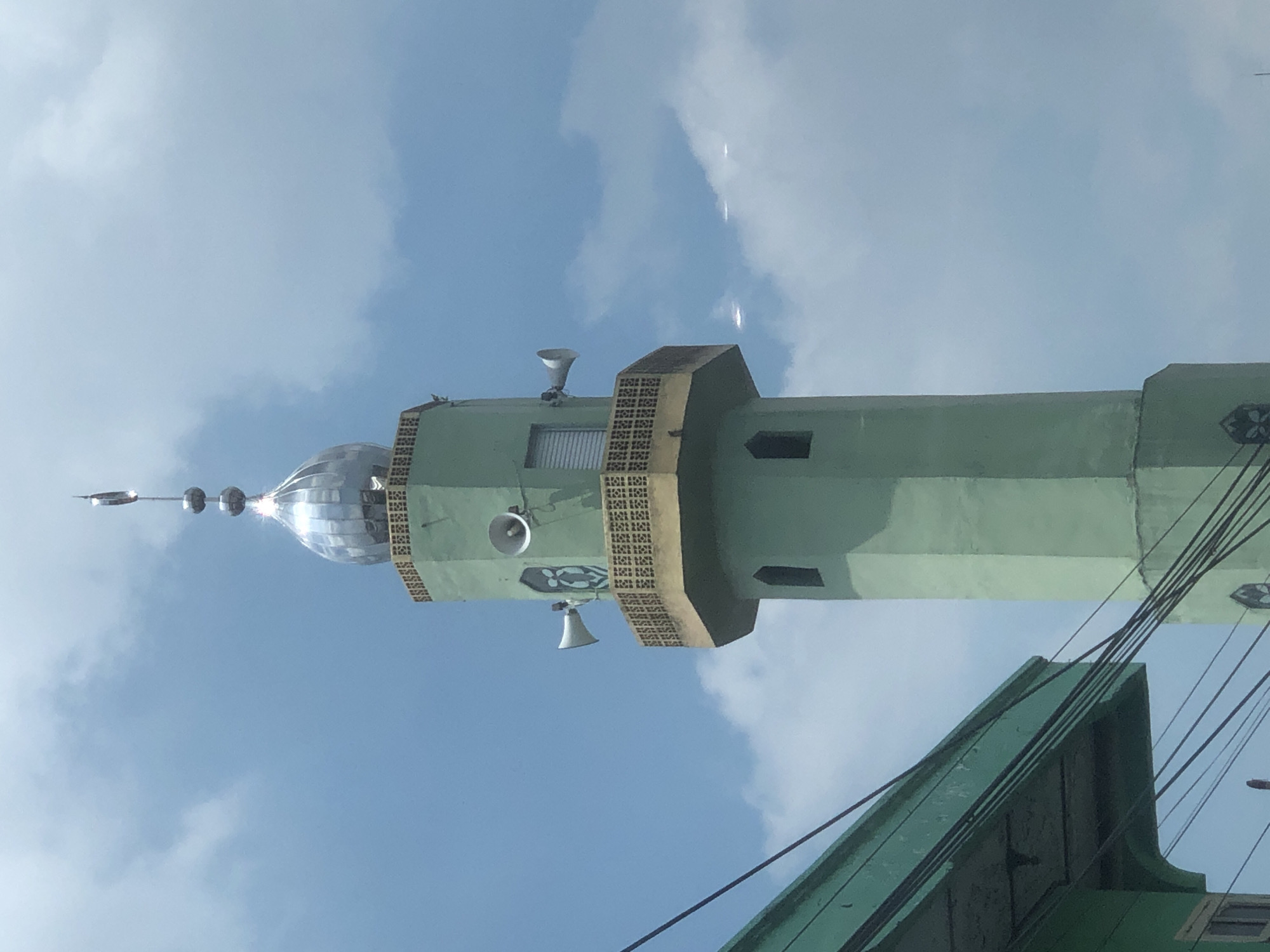 A low-angle, vertically-oriented photograph depicts the minaret of a mosque against a backdrop of a sky with some clouds. The minaret is painted in shades of green, with a silver dome atop. The dome features a crescent moon and several spherical elements. The minaret is embellished with a patterned band and what appear to be loudspeakers. The corner of a green building is visible in the lower portion of the frame. Power lines cut across the lower right. The location is likely Medan, Indonesia.