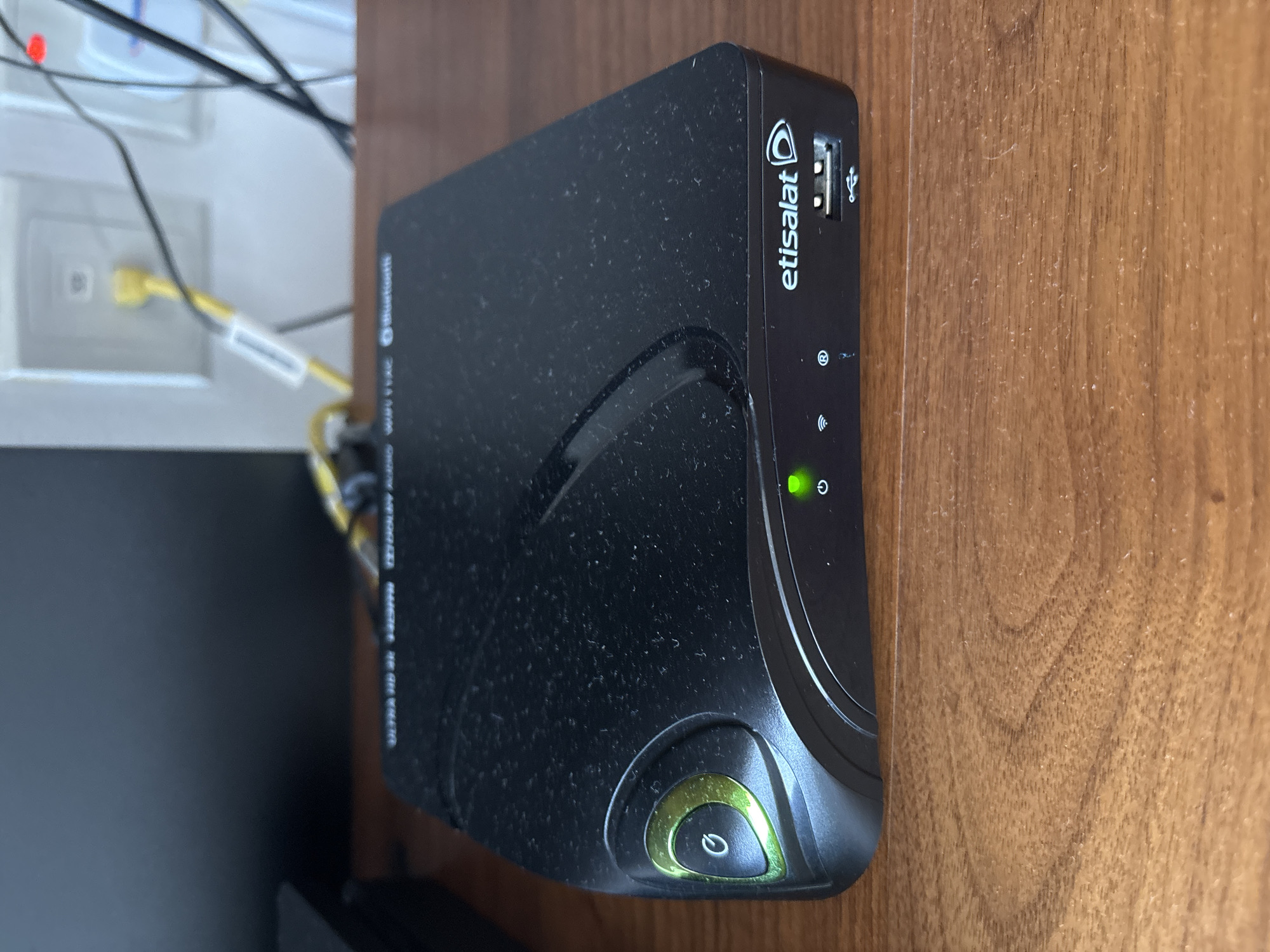 A black Etisalat set-top box is situated on a wooden surface. The device has a green power button on the left side and indicator lights and a USB port on the front. Text on the device reads "ULTRA HD 4K HDR" and "etisalat". The background shows a wall outlet with a yellow cable plugged into it. This scene is likely in Dubai, United Arab Emirates, given the branding of the device.