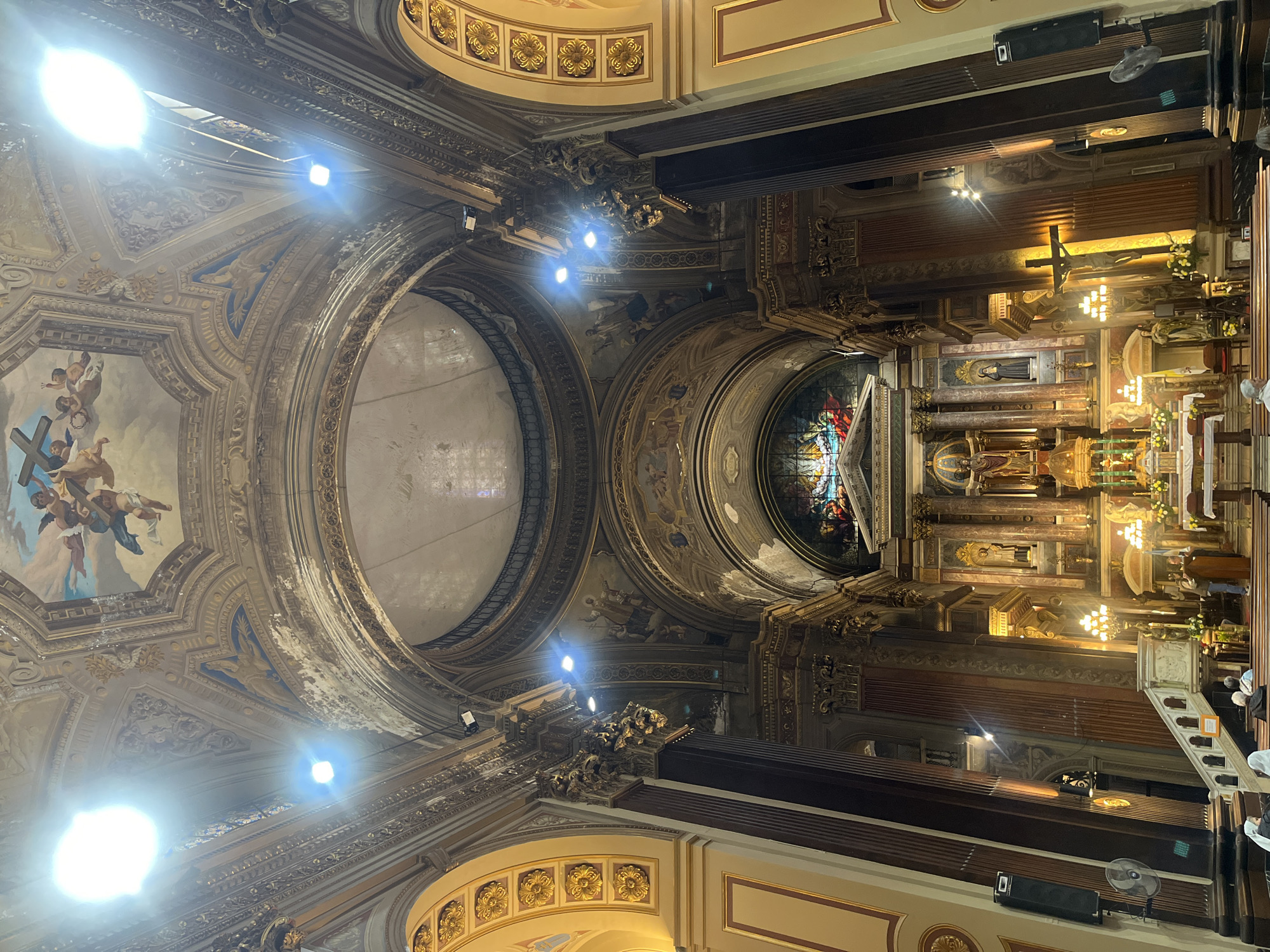 View inside the Catedral Metropolitana de Buenos Aires, Argentina, showing the ceiling and altar. The ceiling is adorned with paintings, gold leaf, and architectural details. The altar is decorated with statues, candles, and flowers. People are sitting in pews. The environment is dimly lit.
