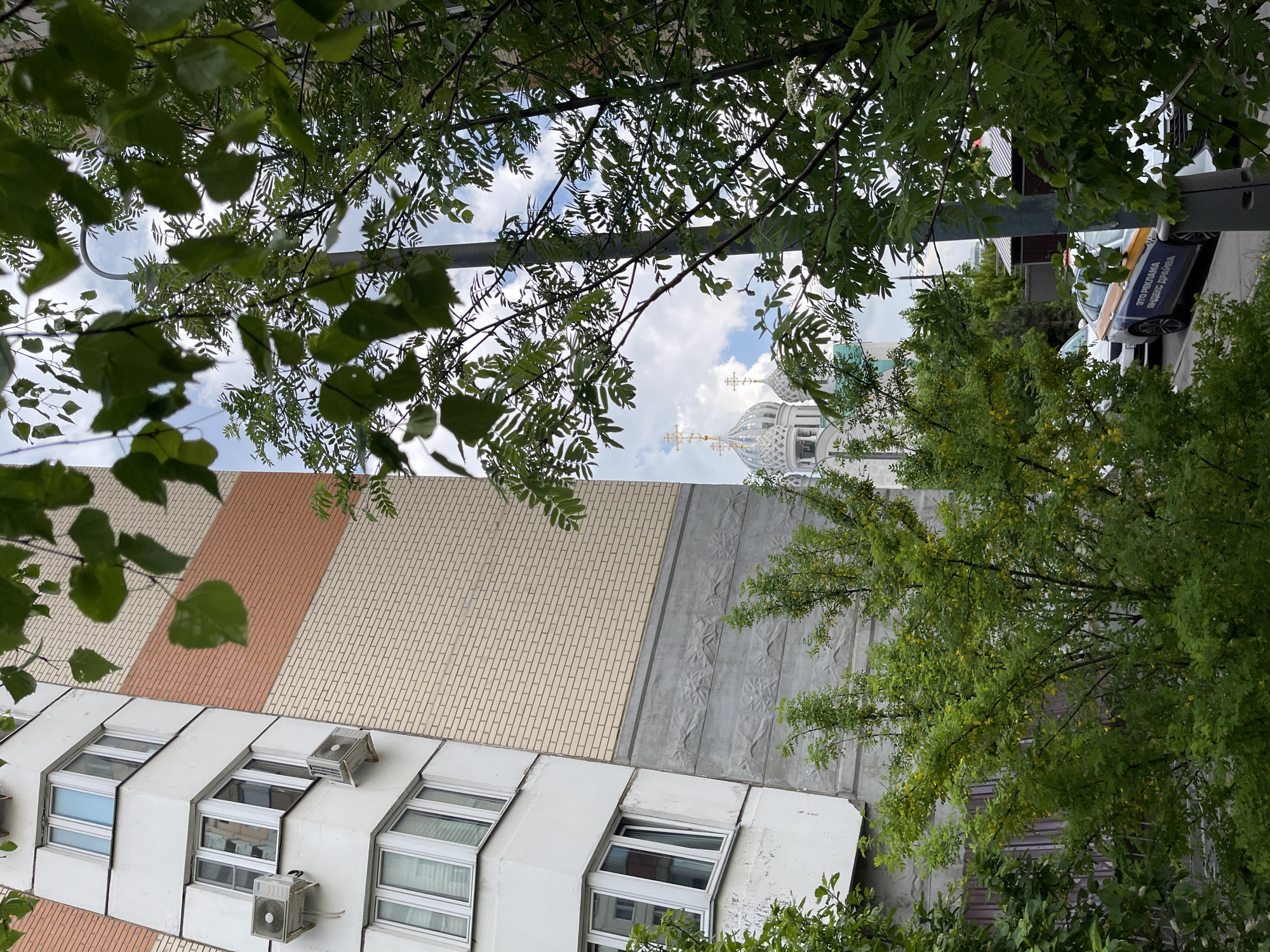 The photograph, taken from a low angle, captures a view of buildings and vegetation in Khimki, Russia. The foreground is filled with green foliage, obscuring parts of the scene. Visible buildings include a multi-story structure with a patterned facade of white, beige, and brown brick, featuring windows and air conditioning units. Through a break in the trees, the upper portion of a Russian Orthodox church, identified by its white dome and golden cross, is seen against a partly cloudy sky. A parked vehicle is partly visible, with text that reads "ЭТО РЕКЛАМА ЯНДЕКС ДРАЙВA" ("This is an ad for Yandex Drive"). A vertical metal pole is also present.