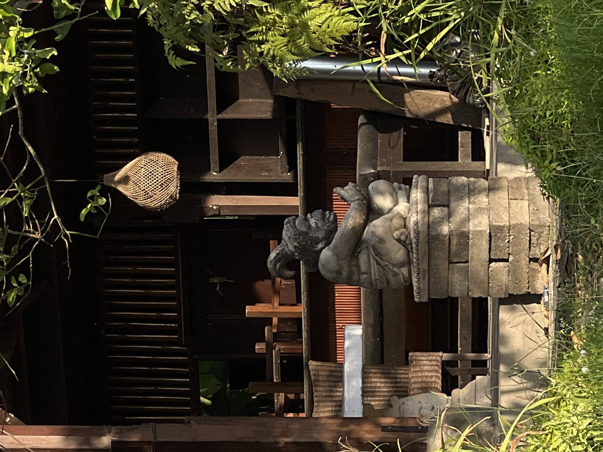 The image depicts an outdoor scene in Mengwi, Indonesia. A gray stone statue of a seated figure is prominently displayed. To the left of the statue, there's a dark, open structure with wooden shelving and a woven lamp hanging. The foreground includes dark wooden elements. Lush green foliage frames the scene on both sides, adding a natural touch to the urban setting. Building materials or architectural fragments are stacked next to the statue.