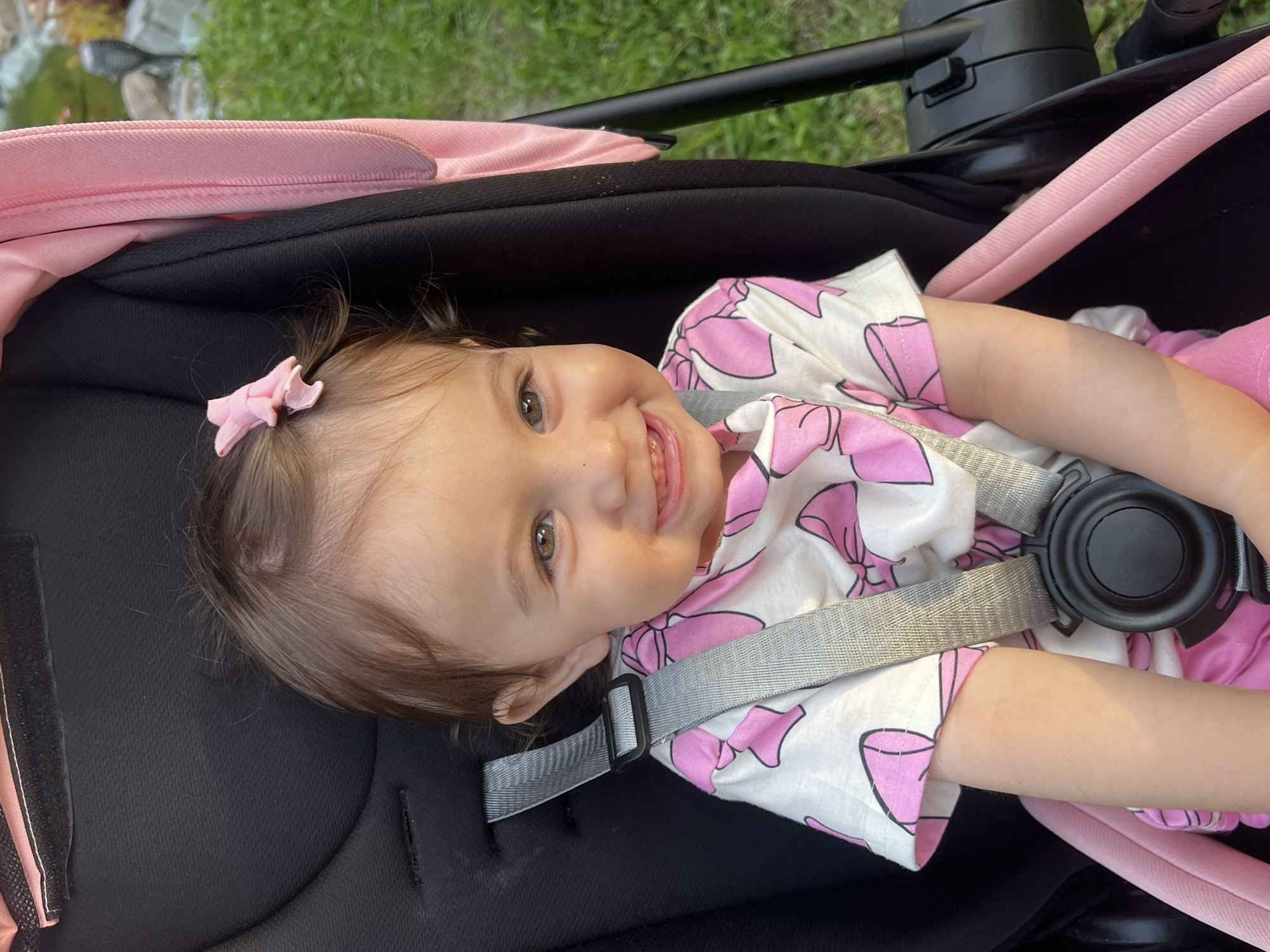 A young child with brown hair and green eyes is sitting in a black stroller with pink trim. The child is wearing a white t-shirt with pink bows and pink shorts. A pink bow is in the child's hair. The stroller is outside on a grassy area with some rocks and a lamppost visible in the background. The image is taken from a close-up perspective, focusing on the child's face and upper body. The sun appears to be setting, casting a warm glow on the scene. No specific location in Elena, Bulgaria, can be identified from the image.