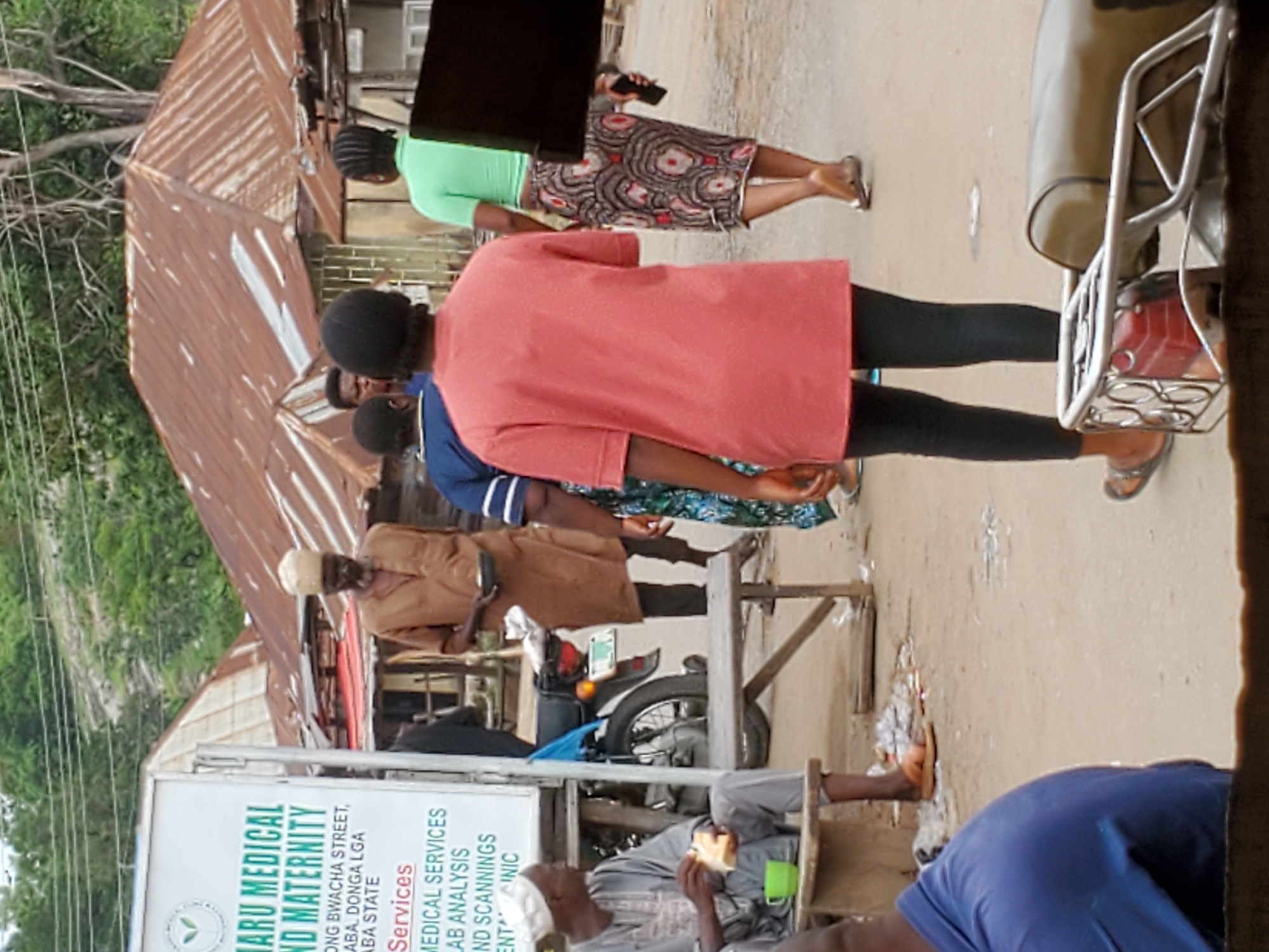 The image depicts a street scene. A sign reading "Maru Medical and Maternity" is visible. People are present, some standing and walking. A man sits eating. Buildings are in the background. A motorcycle is present. Trees are visible in the distance. The OCR text from the sign indicates the location is in Mararaba, Donga LGA, Taraba State, Nigeria.