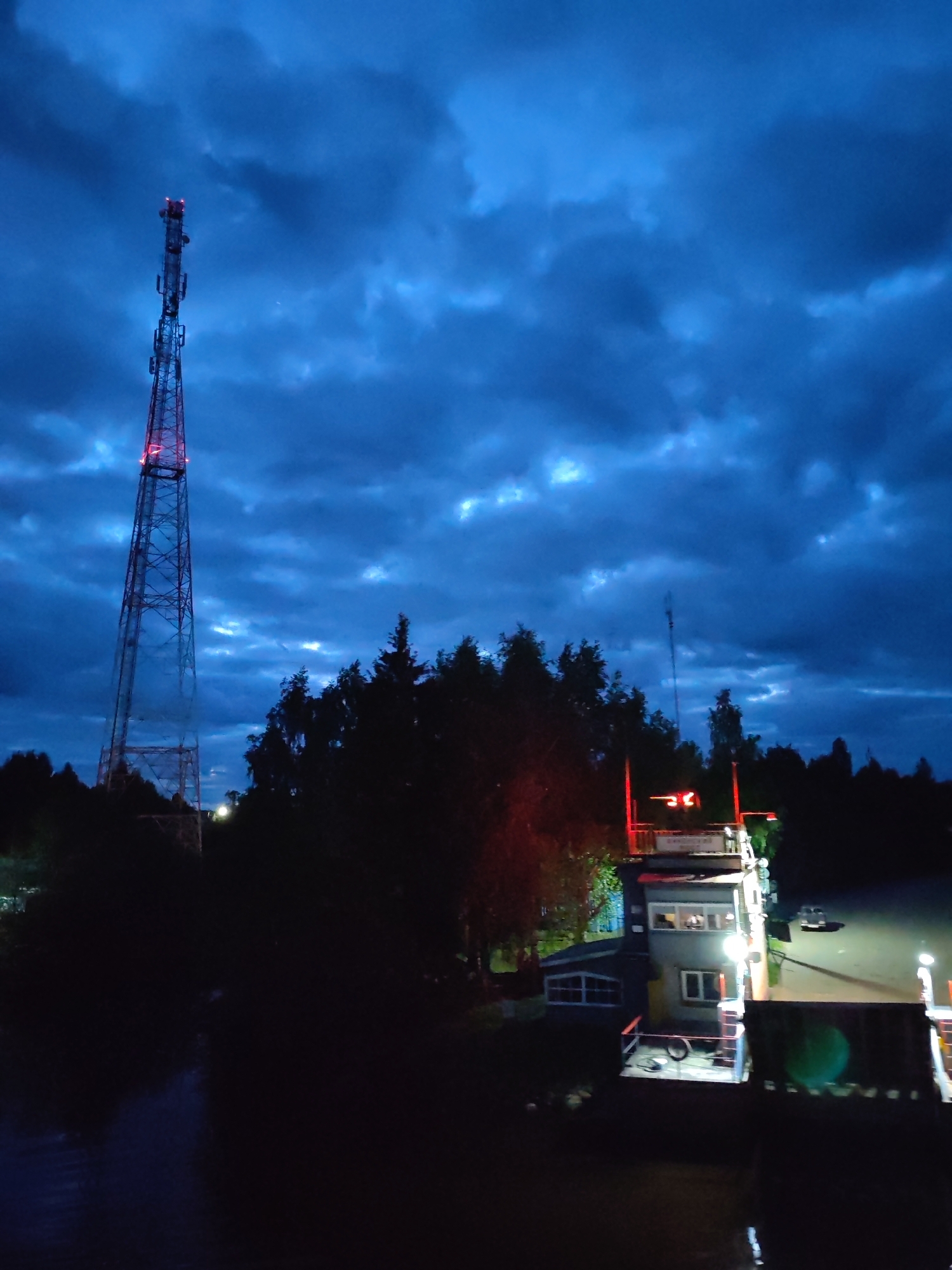 Observed at night in Annensky Most, Russia, a tall, latticed communication tower with red marker lights stands prominently against a cloudy sky. Adjacent to the tower are densely packed trees. To the right of the trees is a two-story structure featuring windows and illuminated by artificial lights. A platform extends towards the water, with objects or equipment discernible on the platform, including what appears to be a bicycle tire. The environment consists of a dark body of water in the foreground. The scene is dominated by dim lighting and dark tones, with a sense of stillness.