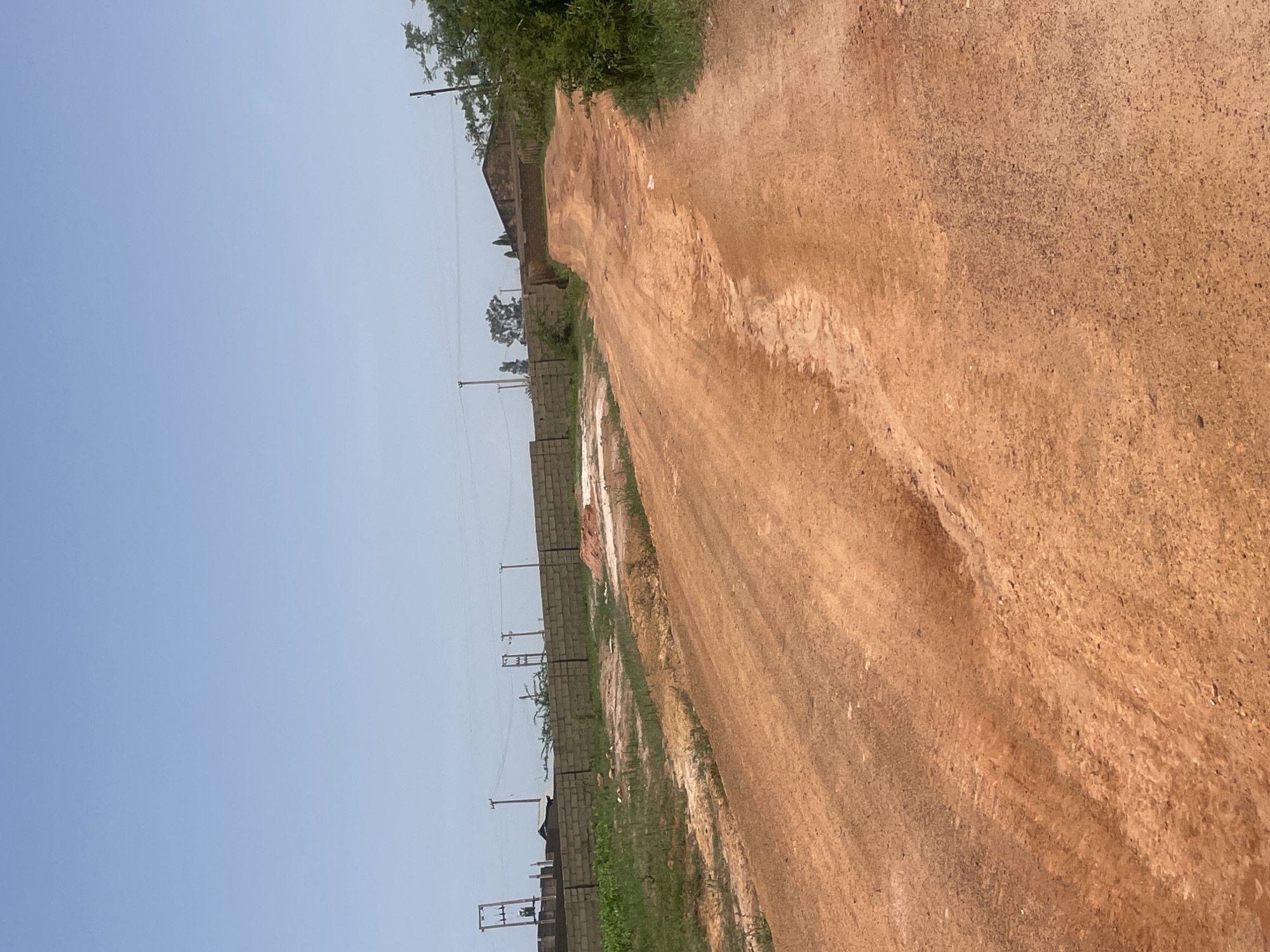The image depicts an unpaved, reddish-brown dirt road that winds upwards and away from the viewer. The road surface shows signs of wear, with tire tracks and some unevenness. To the left of the road, there is a patch of dry, sparse vegetation and a low brick wall. Beyond the wall, more construction materials, including what appear to be stacked bricks, are visible, along with electrical poles and wires. In the background, there is a long brick wall extending horizontally across the frame, with some trees and buildings visible behind it. To the right of the road, there is a dense growth of green foliage, suggesting trees and bushes. The sky above is clear and pale blue, indicating an outdoor scene under daylight. The setting appears to be on the outskirts of Jos, Nigeria, likely an area undergoing development or construction. There are no people or animals present in the image.