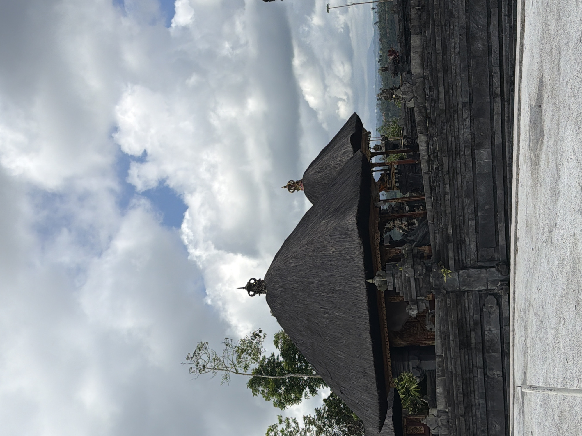 A temple with thatched roofs and ornate stonework is shown under a cloudy sky in Petang, Indonesia. The temple features dark stone walls and steps leading up to the buildings. Two conical, thatched roofs are prominent, each topped with a decorative crown. Behind the temple, lush green trees and distant hills are visible. The foreground consists of a light-colored, possibly concrete, surface. The image captures a traditional architectural style against a natural, expansive backdrop.