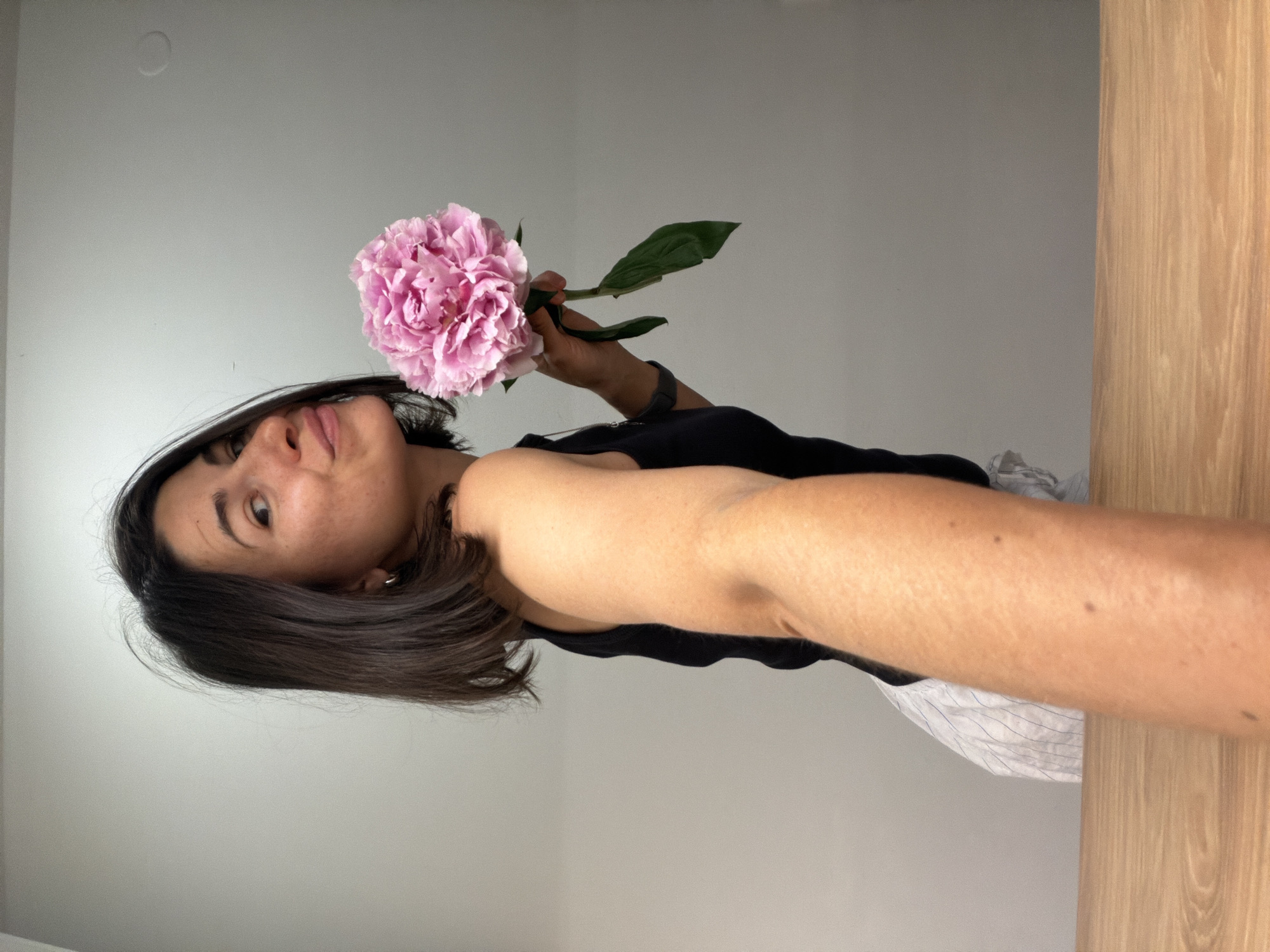 A woman poses holding a pink flower with green leaves. She is wearing a black tank top and a white garment with blue stripes. Her dark hair is shoulder-length. The background consists of a light gray wall and a light brown wooden surface. There is no indication of the city of Budva, Montenegro.