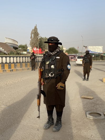 Three armed individuals are standing on a city street in Kandahar, Afghanistan. All three wear brown uniforms, hats, and face coverings. The individual in the foreground holds a rifle. A white vehicle is in the background. A road divider and buildings are in the background. The weather appears sunny and clear.