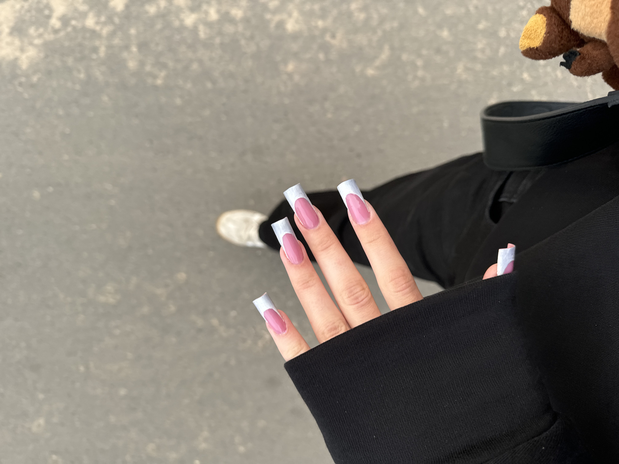 A person's hand with pink and light blue french tip acrylic nails is positioned in the foreground, partially covered by a black garment. The person is wearing black pants and white sneakers, suggesting they are outdoors on a paved surface, possibly a street or sidewalk. A portion of a black bag and a brown plush toy are visible to the right. The background consists of a grey, mottled surface, likely asphalt or concrete. The lighting suggests it is daytime. The image does not provide any specific indicators to determine its location within the city of Сароцкі сельскі Савет, Belarus.