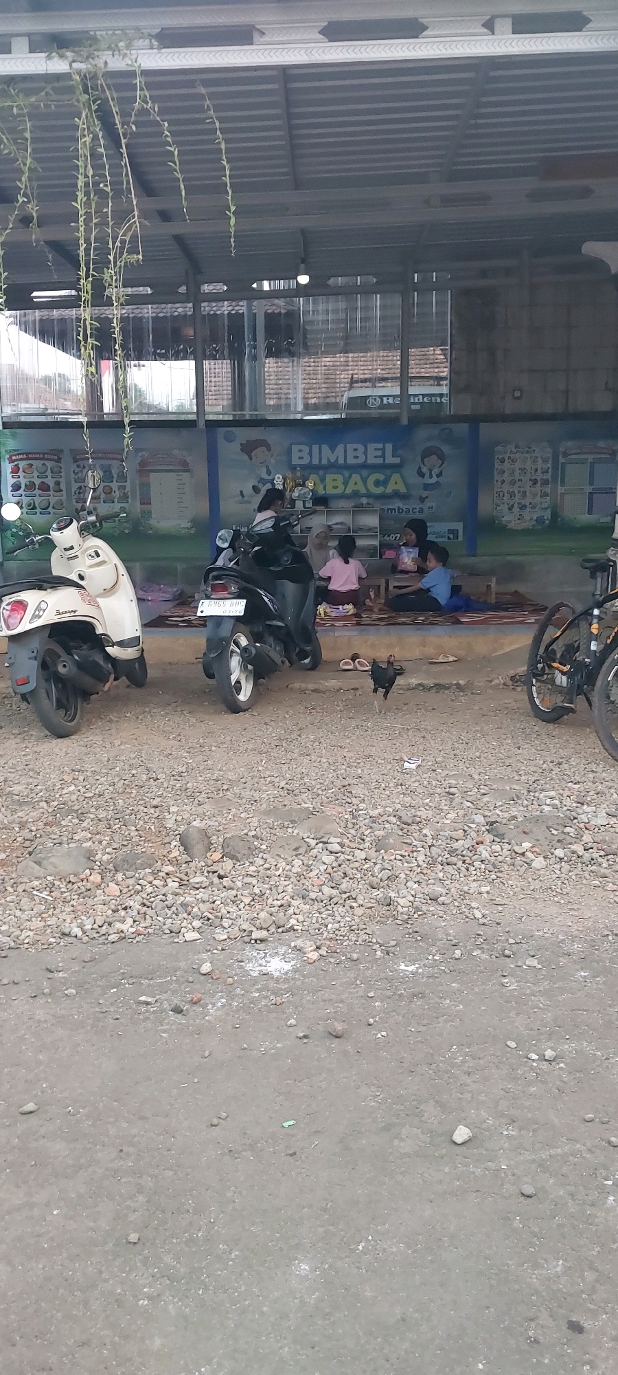 The image shows a side view of a Bimbel (private tutoring) center in Keling, Indonesia. The center is under a metal awning, with a blue mural depicting children and the words "BIMBEL BACA" (Reading Tutoring). Inside the open-air center, several children and an adult are seated on the floor, engaged in activities. Two scooters are parked in front of the center, one cream-colored and the other black. A chicken is walking on the gravelly ground in front of the scooters. In the background, a bicycle and more scooters are visible. The ground in front of the center is covered in gravel and dirt.