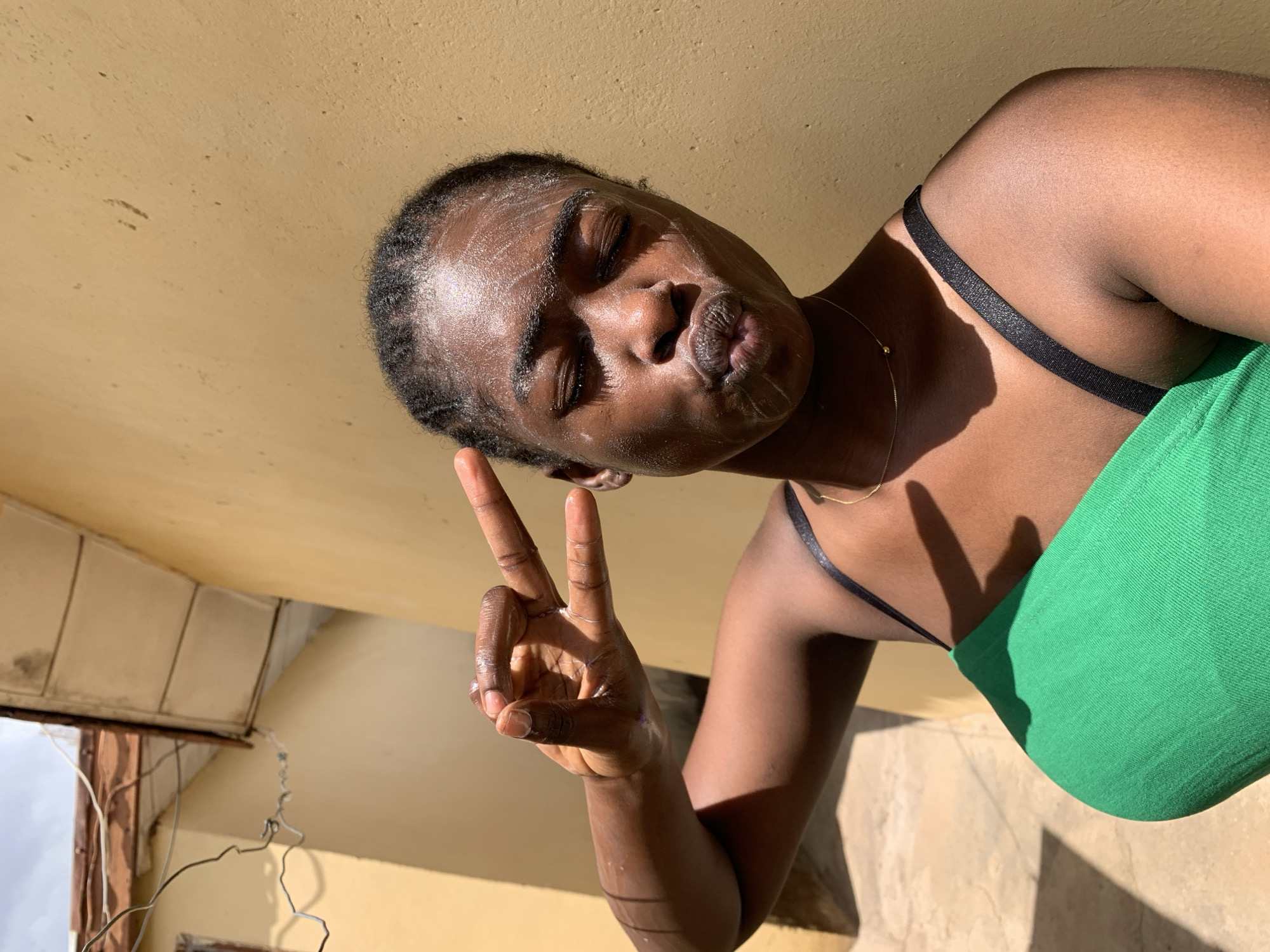 A person with dark skin is posing for a selfie in an indoor location, likely Rafin Par, Nigeria. The person is wearing a green top with black straps and has a necklace with a small gold bead. Their face appears to have a white, creamy substance, possibly a face mask. The person's eyes are closed, and they are pouting their lips while holding up a "peace" sign with their fingers. The background shows a yellowish ceiling and the corner of a window or door frame. Electrical wires are also visible near the frame. Sunlight is casting shadows on the person and the surfaces.