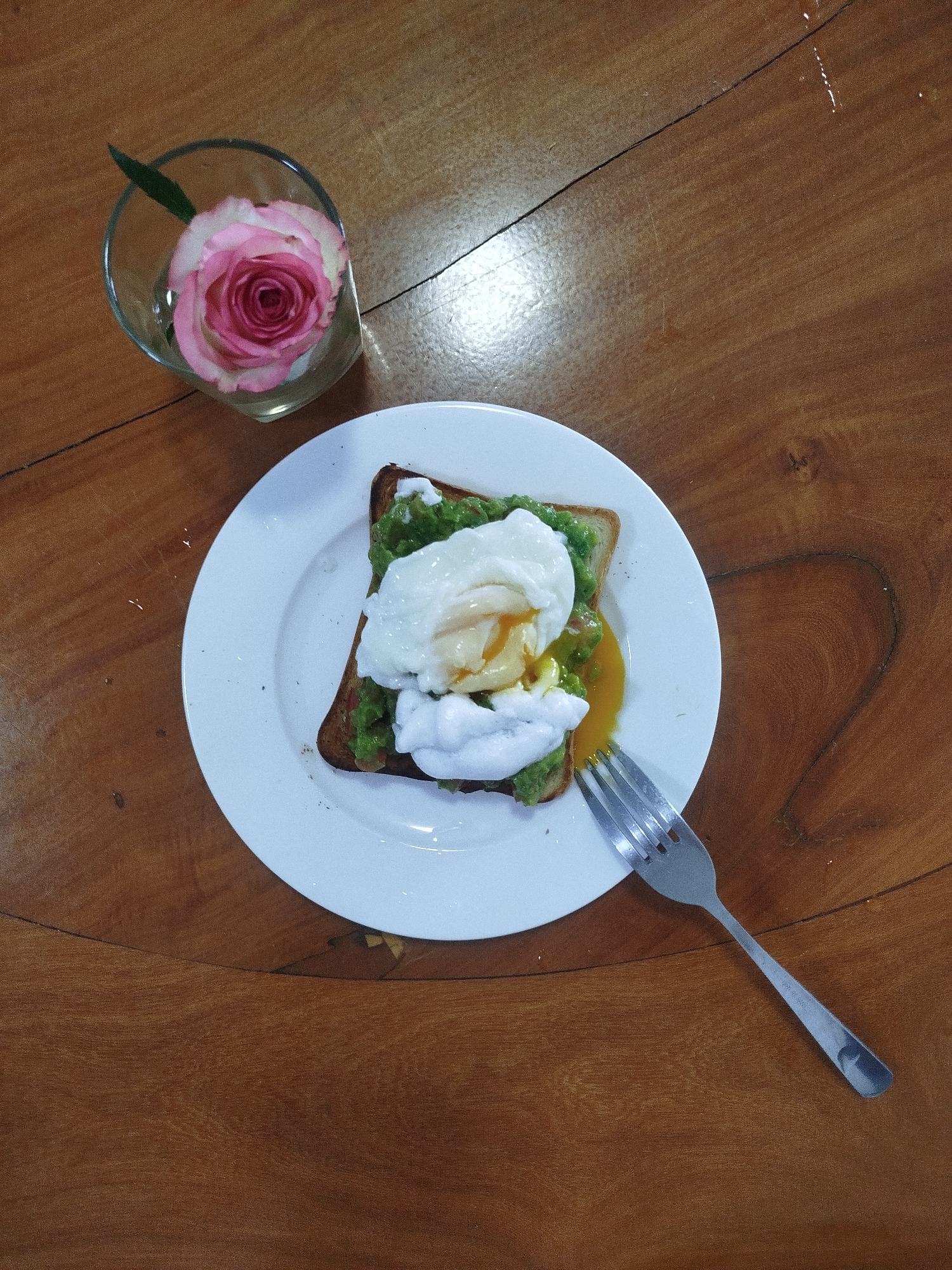 The image shows a plate of avocado toast topped with a poached egg and a fork on a wooden table. Next to the plate, there is a small glass with a pink rose inside. The scene is likely inside a cafe or restaurant in Da Lat, Vietnam, based on the context. There are no visible people. The action is focused on the presentation of the food. The environment is simple and natural, with the wooden table and the soft lighting.