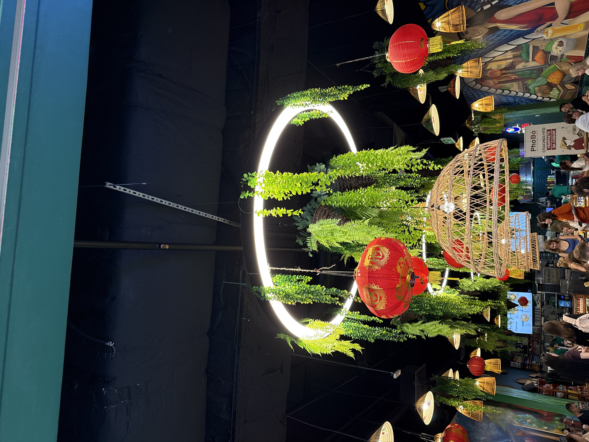 The image depicts the interior of a restaurant, possibly located in Moscow, Russia, based on Cyrillic script on a sign. The space has an eclectic decor with hanging light fixtures, including red paper lanterns and conical lampshades. A prominent circular light installation is adorned with greenery. Patrons are seated at tables, and a mural is visible on the wall. The scene appears to be an active dining area.