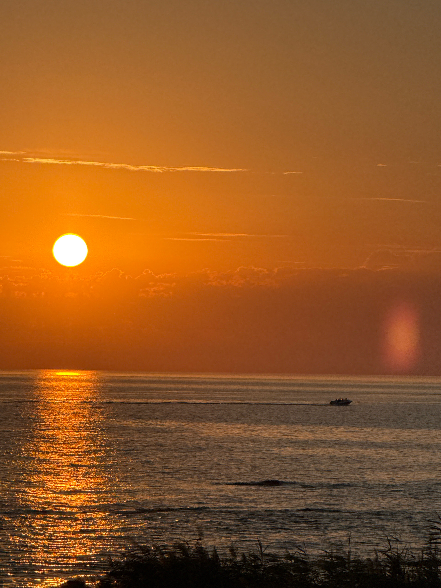 The image depicts a sunset over the Caspian Sea, with the sun appearing as a bright, white orb near the left horizon. The sky is a gradient of warm orange and yellow hues, with thin, wispy clouds scattered across the upper portion. A reflection of the sun shimmers on the calm surface of the sea, creating a bright path of light. In the middle distance, a small boat with several occupants is moving from left to right, leaving a faint wake. In the foreground, dark silhouettes of reeds or tall grass line the bottom edge of the frame. A faint, vertical light phenomenon, possibly a sun pillar, is visible in the right portion of the sky. The overall scene is tranquil and serene, capturing the essence of a coastal sunset. This scene is consistent with the typical sunsets observed along the coast of Aktau, Kazakhstan, which is situated on the Caspian Sea.