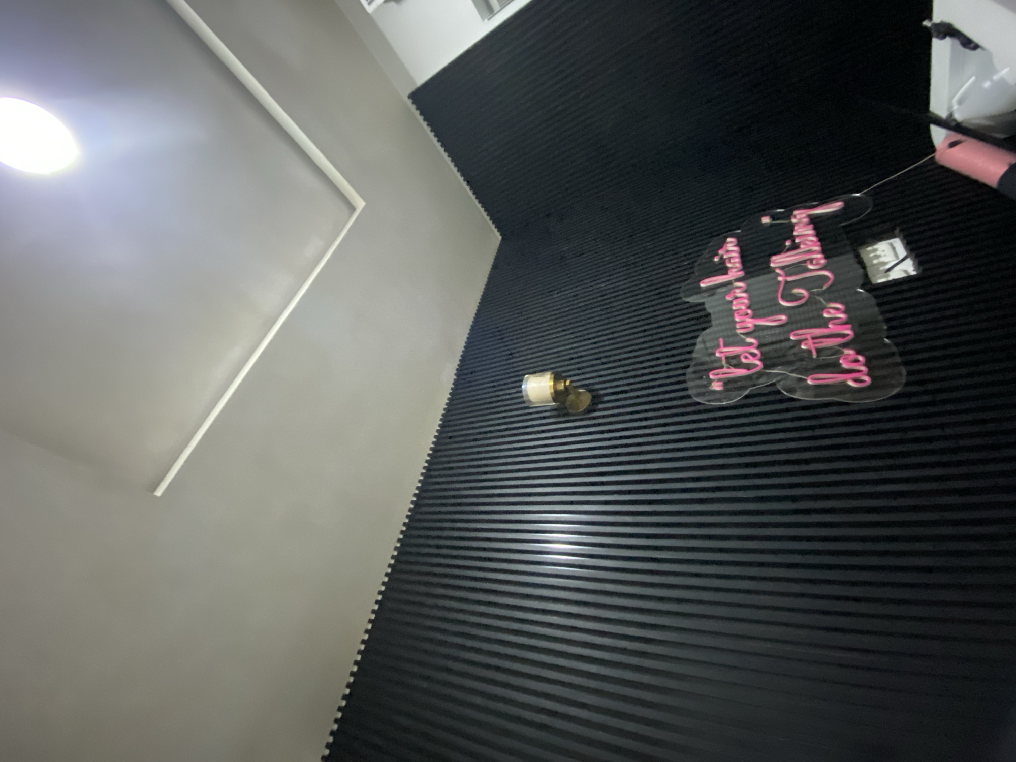 The image depicts an interior space in Lagos, Nigeria, showcasing a room with contrasting wall designs. The left wall is painted in a neutral gray tone, while the adjacent wall is covered in a dark, horizontally-striped pattern, creating a visually textured effect. A lighted sign with pink writing is mounted on the striped wall. Above the sign, there is a power strip. A candle is mounted on the striped wall. A light fixture is visible in the upper-left corner of the image.