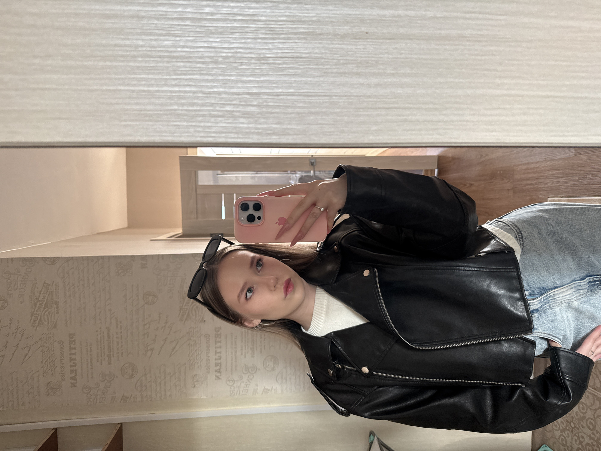 The image shows a young woman taking a selfie in a mirror. She is wearing a black leather jacket over a white sweater and light blue jeans. Sunglasses are perched on her head. She is holding a pink phone, likely an iPhone, in her right hand.
The reflection in the mirror shows a hallway with wallpaper decorated with a patterned print, a closed light-colored door, and a window shade partially lowered.
There is no verifiable information confirming this photo was taken in Chaikovsky, Russia.