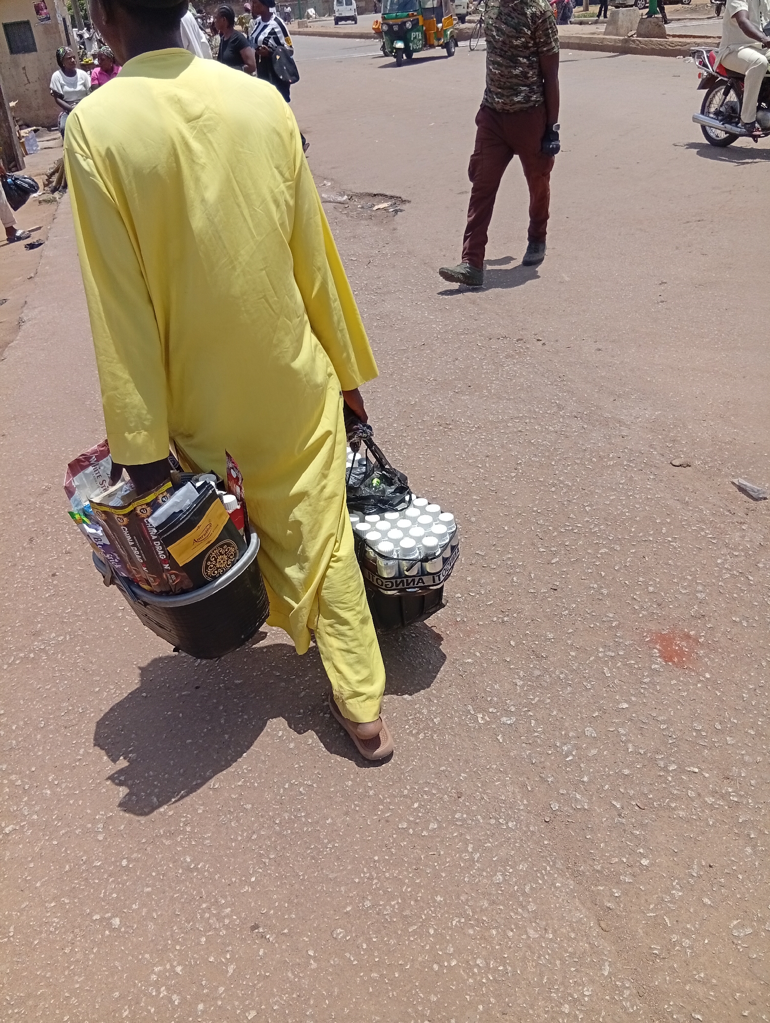 A man in a yellow robe is carrying two black buckets through the streets of Bukuru, Nigeria. One bucket is filled with packaged foods, and the other with small, capped bottles. Another man in camouflage and brown pants is walking in the same direction. Other people are visible in the background. Vehicles, including a tricycle and motorcycles, are also in the background. The ground is paved.