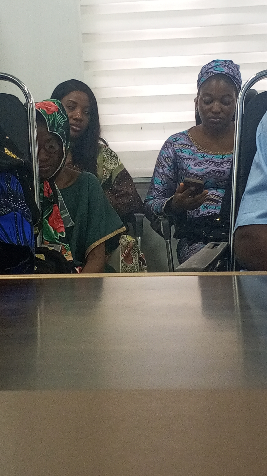 The image captures an indoor scene, likely a meeting room, classroom, or office, with at least four individuals, predominantly women, partially visible. In the foreground, a smooth, light brown table surface occupies the lower third of the frame. Behind the table, several people are seated, their upper bodies mostly visible.
On the left, a woman with dark skin wears glasses and a colorful headscarf featuring red roses on a green background. She is dressed in a green top over a patterned garment and appears to be looking downwards or to the left. Behind her, another woman with dark skin and long dark hair looks towards the right with a neutral expression, wearing a patterned top with earthy tones.
To the right, a woman with dark skin is prominently featured, wearing a patterned blue and purple head covering and a matching long-sleeved dress. She is actively engaged with a smartphone held in both hands, looking down at the screen. Another smartphone, encased in grey, rests horizontally on a surface in front of her. Partially visible on the far right is the shoulder and arm of a person in a light blue shirt.
Metallic chair frames are visible, indicating seating. The background consists of horizontal white blinds, suggesting a window or partition. The overall expressions are neutral, and the lighting is consistent with an indoor setting. There is no visible text in the image. The attire, particularly the head coverings and patterned garments, suggests a cultural context consistent with Lokoja, Nigeria.