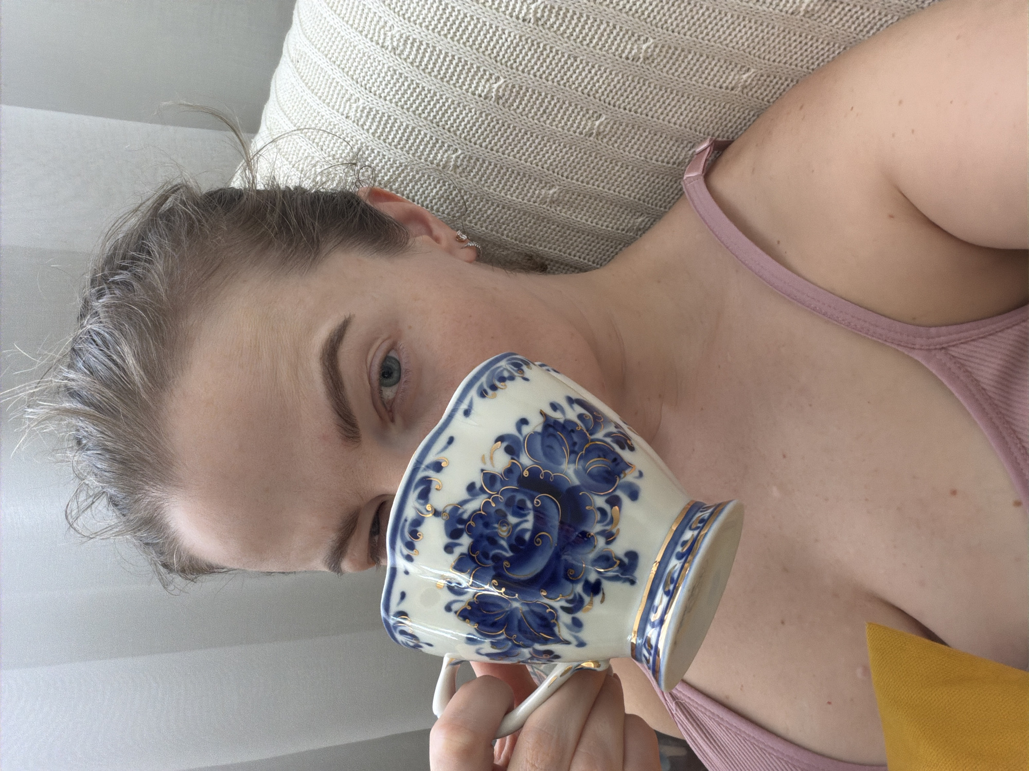 A fair-skinned person is lying on a textured light-colored pillow, holding a white and blue Gzhel-style teacup. They wear a pink tank top. The scene takes place indoors, possibly in Moscow, Russia, given the style of the teacup.