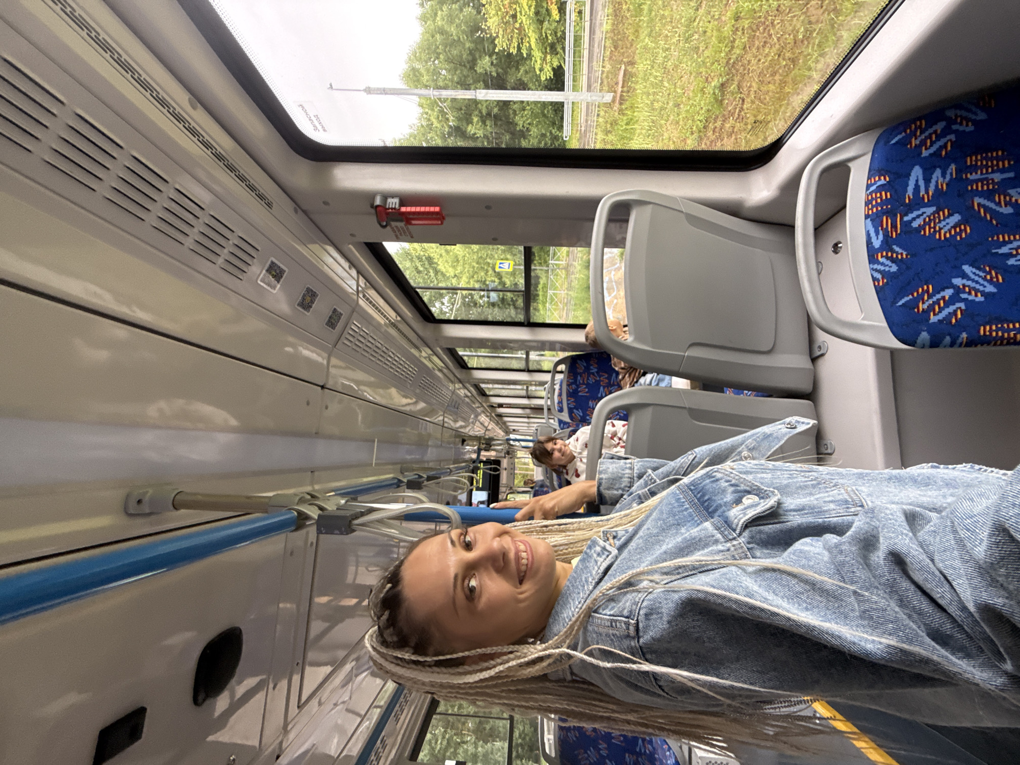 A blonde woman with braided hair smiles and poses for a photo on a bus. She is wearing a denim jacket. Other passengers are visible in the background, and the bus is moving through a green, wooded area. The location is identified as Rusanovo, Russia.