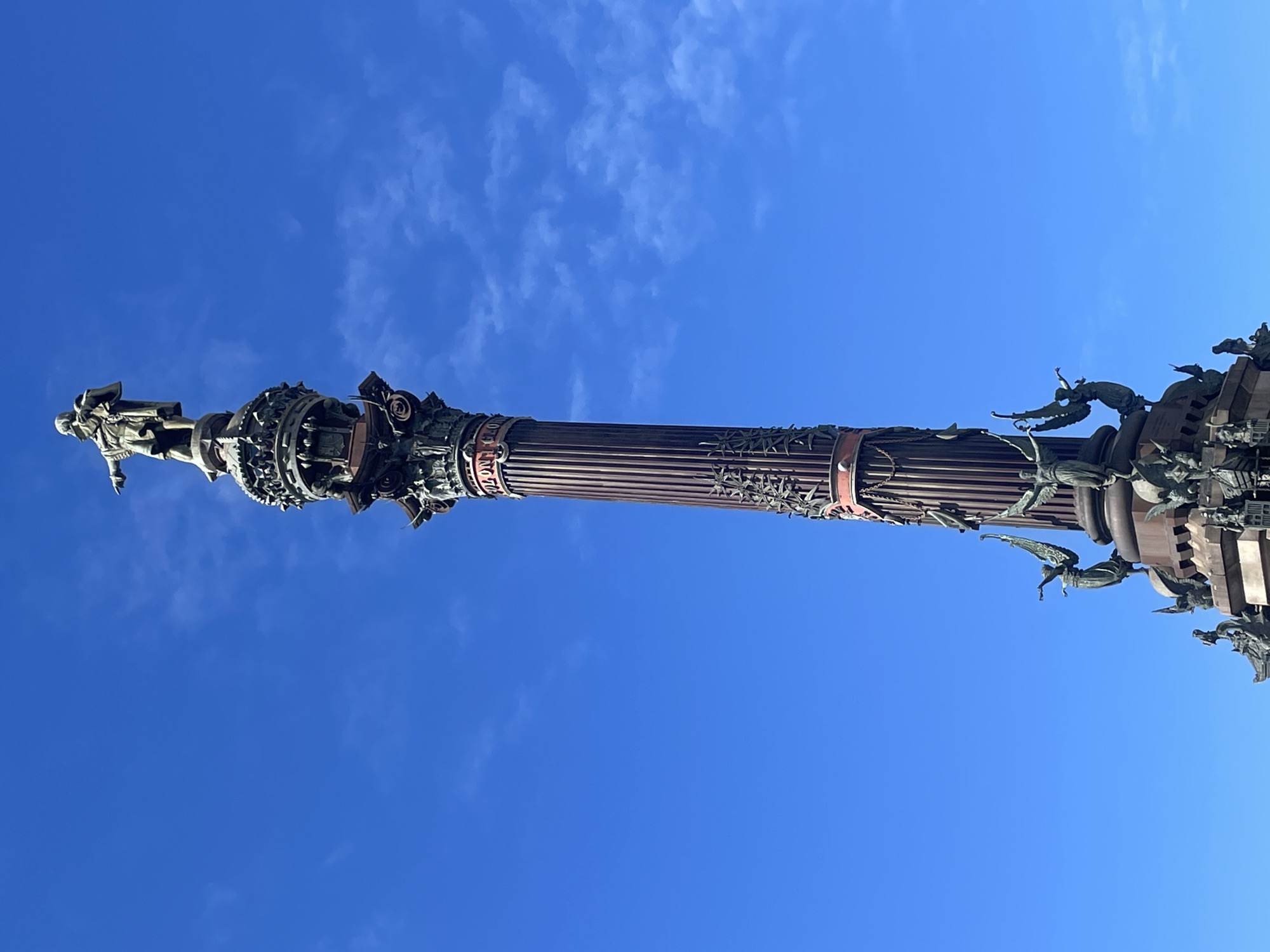 The image shows the Columbus Monument in Barcelona, Spain, seen from a low angle looking up at the monument against a clear blue sky with some wispy clouds. The monument consists of a tall, fluted column topped with a statue of Christopher Columbus pointing out to sea. The column is decorated with bronze figures and nautical motifs. The base of the monument is obscured by the upward angle.