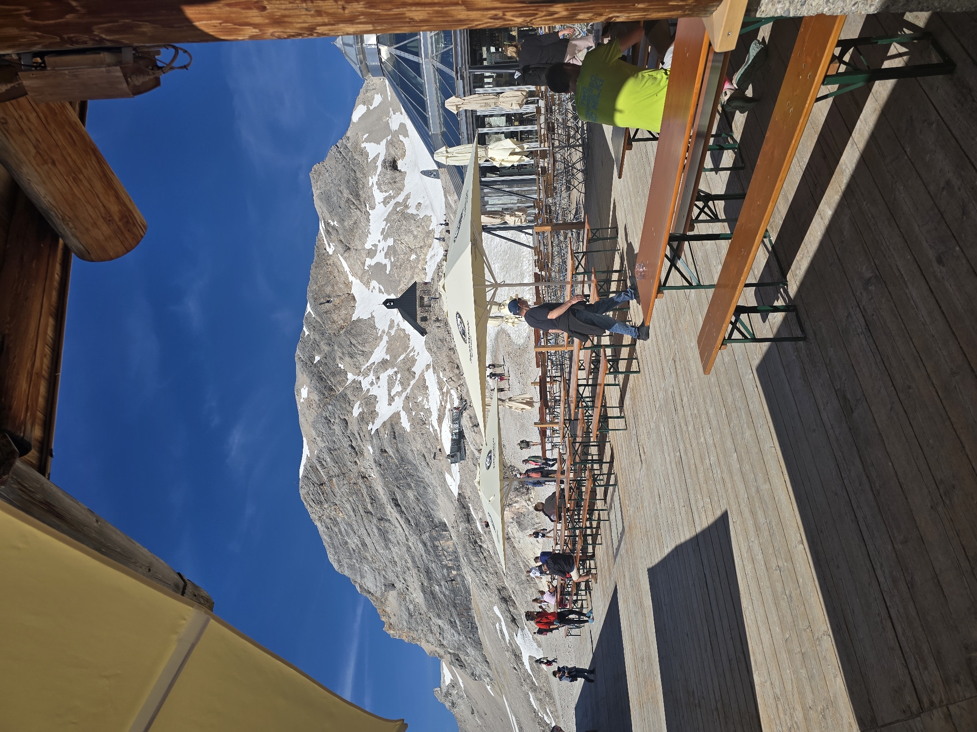 This media file depicts a sunny day at a mountain-top restaurant with a panoramic view of a snow-dusted mountain range. The scene is set on a wooden deck filled with outdoor seating, including long wooden tables and benches, some of which are occupied by people.
Several individuals are visible in the frame, appearing relaxed and enjoying the scenery. One person in a bright green t-shirt is seated at a table, while others are seen walking around or sitting on benches. In the background, the imposing mountain, partially covered in snow, dominates the landscape under a clear blue sky. Structures resembling a building with a glass facade and a dark-roofed smaller building are also present on the mountainside. A yellow awning is visible in the foreground, adding a touch of color to the scene. The shadows cast by the tables and benches suggest it is mid-day. The overall atmosphere appears to be one of leisure and appreciation of the natural beauty of the location. There is no visible text in the image. The location is likely a tourist attraction given the presence of a well-maintained deck and restaurant facilities at a high altitude.