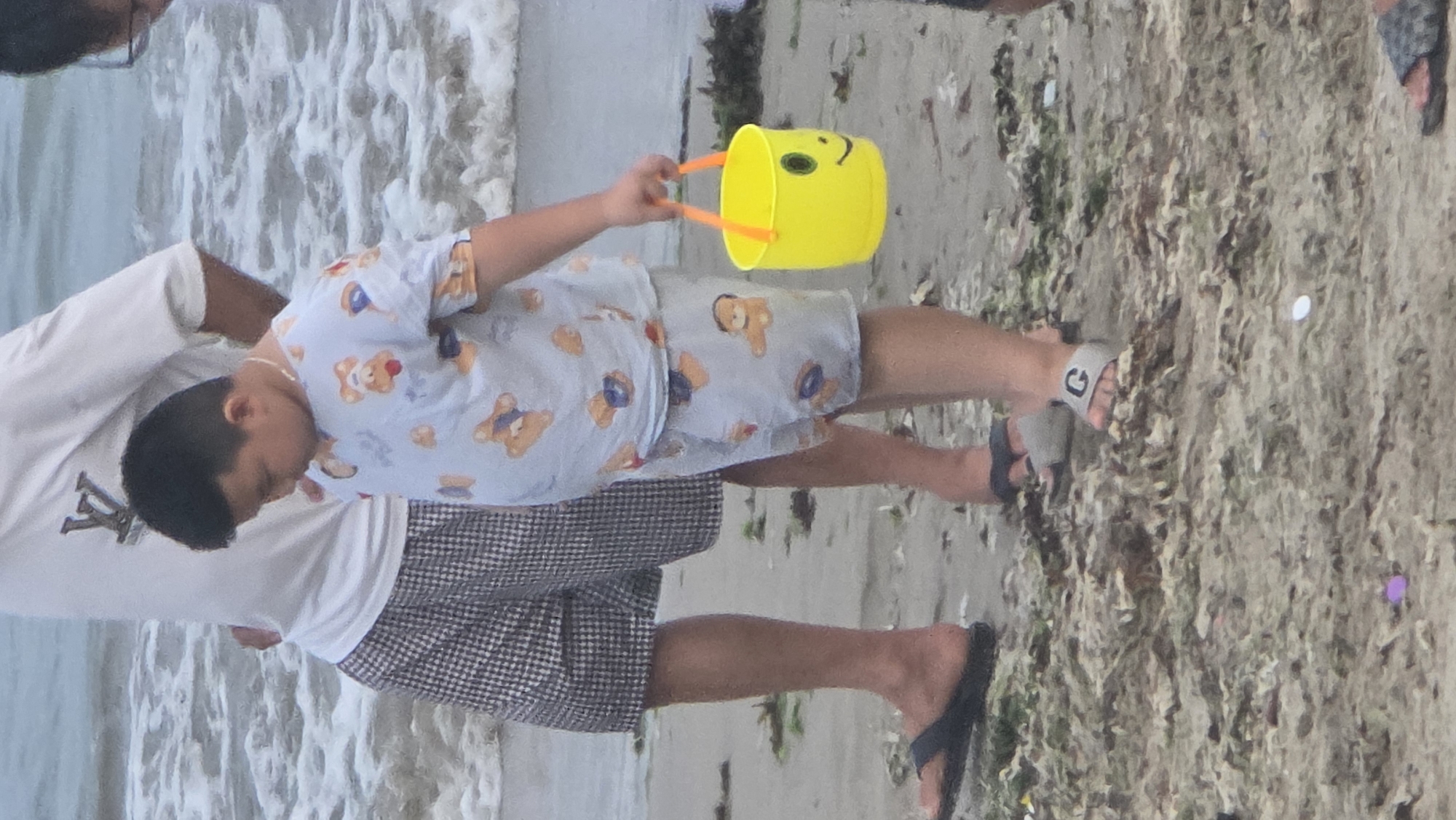 The image depicts a child holding a yellow bucket with a smiley face on a beach. An adult is standing beside the child. The location is likely the beach in the city of Bắc Nha Trang, Vietnam. The beach appears sandy and has some seaweed and small pieces of trash on it. The water is in the background. The child is wearing a light blue outfit with a teddy bear print and sandals. The adult is wearing a white t-shirt with a logo and plaid shorts, along with flip-flops.