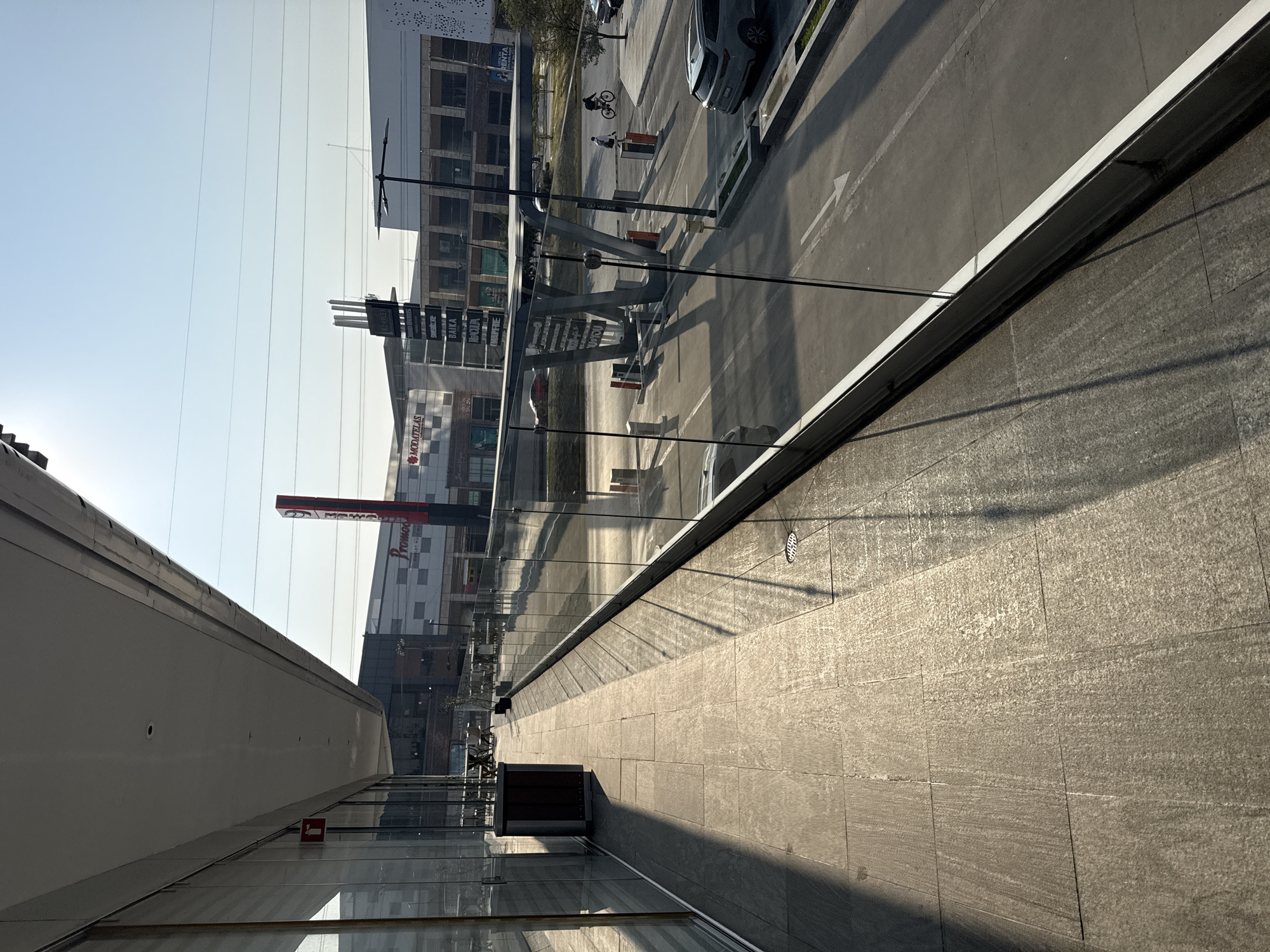 A long-shot, low-angle perspective, taken from an elevated position, oriented vertically, portrays an urban scene at the Explanada Puebla shopping center in Puebla, Mexico. A view outside, encompassing the street, pedestrian areas, and reflections off a glass facade of the center.
In the external view, pedestrians and cyclists traverse the sidewalks, and vehicles are parked or in motion on the road. The retail establishment "PROMODA" and other commercial signage are visible.
Reflections of the external scene are prominent on the glass facade, intermingling with the interior elements, such as the stone tiled flooring.
The light casts shadows across the tiled surface, which suggests either early morning or late afternoon. The weather appears clear and bright.