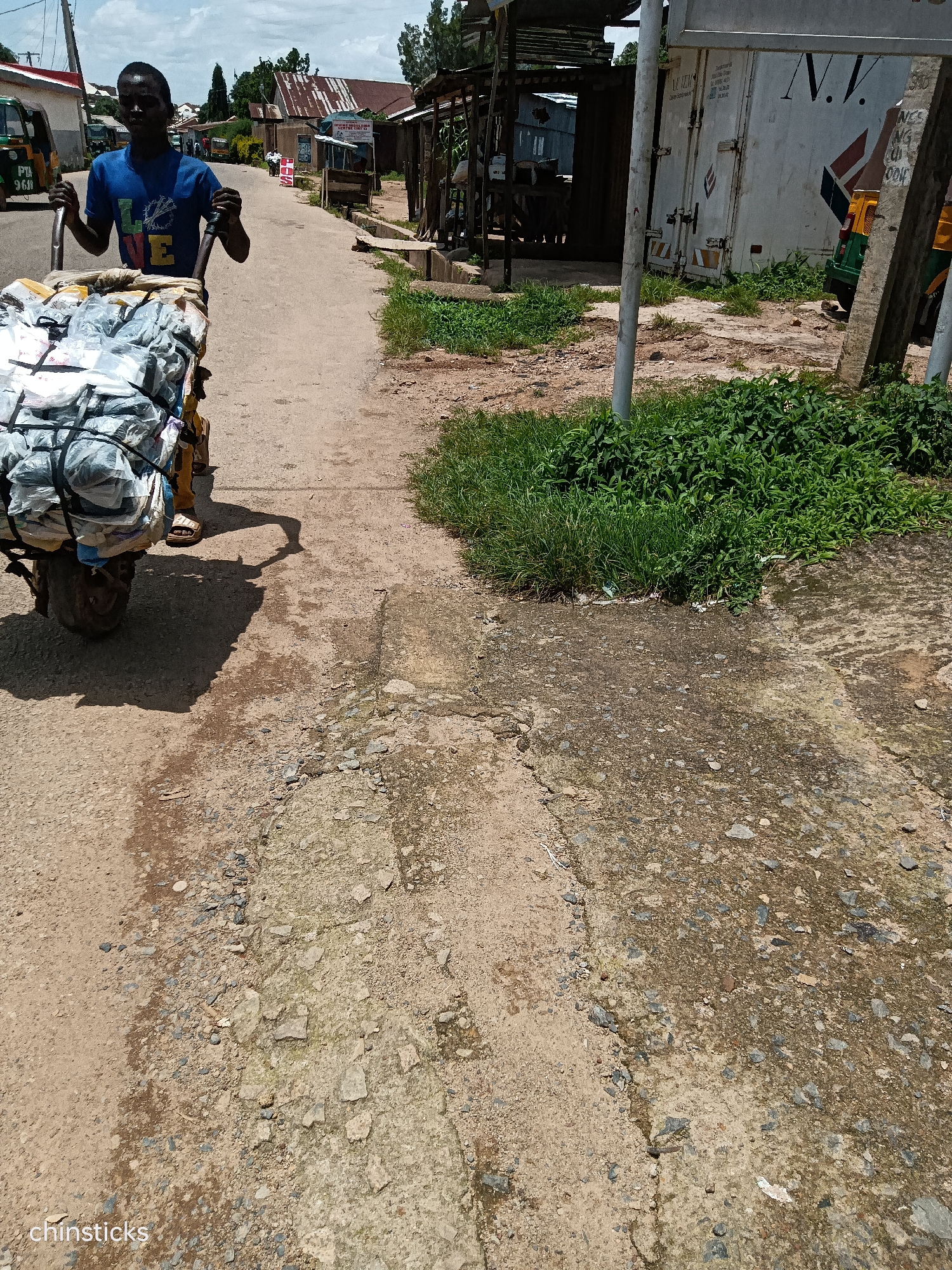 A young Black man is pushing a cart loaded with plastic bags and other assorted items down a dusty, cracked paved road in Jos, Nigeria. The man is wearing a blue t-shirt with a colorful "LOVE" graphic and dark shorts. He is pushing the cart with both hands on the handles, his body leaning forward. The cart has two wheels and is laden with a large volume of white and gray plastic bags, some secured with black straps. The road surface is uneven, with patches of concrete and gravel. On the right side of the road, there is dense green vegetation. In the background, there are buildings, including one with a corrugated metal roof and another that appears to be a shipping container with "N.V." written on it. Several vehicles, including what appears to be a tricycle taxi, are visible in the distance. The sky is partly cloudy. The overall scene depicts a person engaged in the activity of transporting goods in a bustling urban environment.