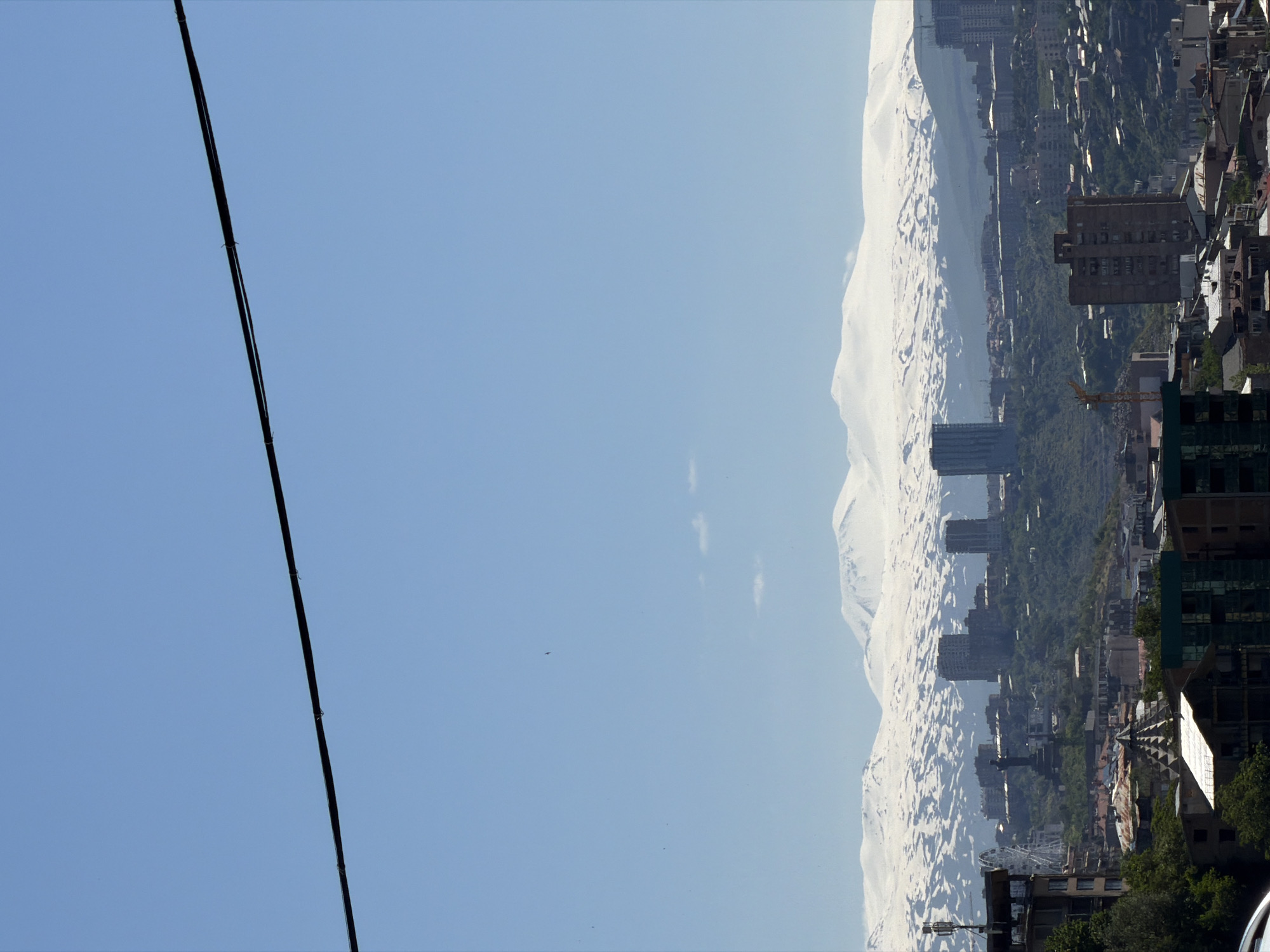 The scene is a landscape view of Yerevan, Armenia, possibly taken from an elevated position. A city with buildings and a ferris wheel is visible. In the background is a snow-capped mountain. A black power line is visible in the foreground.