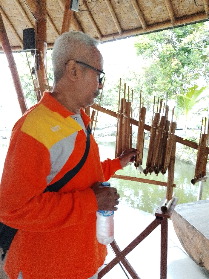 The image depicts an adult male in a semi-outdoor, open-air structure in Pabelan, Indonesia. The man, with short gray hair, a mustache, and dark-rimmed glasses, is shown in right profile, wearing an orange long-sleeved polo shirt with yellow and gray diagonal accents, and a black strap crossing his chest. His right hand is extended towards a collection of bamboo Angklung instruments, which are suspended on a wooden frame over a body of water. His left hand holds a clear plastic bottle of "Aqua" brand water with a blue cap. Above, a woven bamboo roof is visible, supported by bamboo poles, with a black speaker mounted on one such pole. The background features dense green foliage, including trees and a palm frond, beyond the water. A large, light-colored wooden object is partially visible on the lower right foreground.