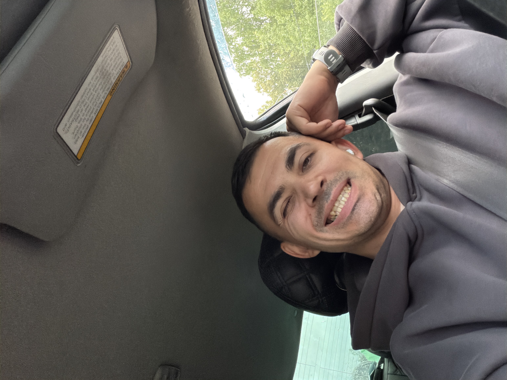 A man is taking a selfie inside a car. He is wearing a grey hoodie and has a watch on his left wrist. The man is winking and smiling, with his hand resting on his head. The background shows trees and foliage outside the car window. The sun visor is visible on the top of the car, with a warning sticker on it. There is no indication of the location being Izhevsk, Russia.