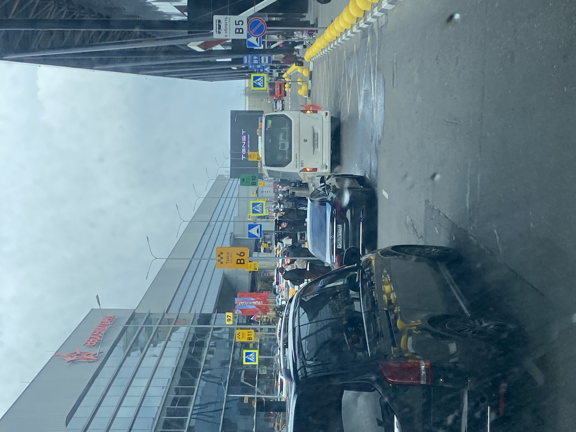 This media file captures a busy outdoor scene, likely at an airport or large transportation hub in Khimki, Russia, from the perspective of a passenger inside a vehicle. The image is taken during the daytime under an overcast sky, with damp-looking asphalt suggesting recent rain or drizzle.
The scene shows multiple lanes of traffic alongside a modern, glass-paneled building on the left, possibly an airport terminal, featuring a large red logo that reads "ПОБЕДА!" (Pobeda!), a Russian low-cost airline. On the right, a structure with yellow protective bollards lines a multi-lane road where vehicles are either stopped or moving slowly.
Visible subjects include several cars, mostly dark-colored sedans and SUVs, and a prominent white passenger van with "881" on its rear, which appears to be a shuttle or taxi. A license plate "Y562KT792" is visible on one of the cars. Numerous pedestrians are seen walking on the sidewalks next to the building and between parked cars, many wearing jackets, indicative of cool weather.
The area is filled with various informational and traffic signs. Prominent signs include "B5" and "B10" with bus icons, some indicating "в область" (to the region). Other yellow signs mark "Такси" (Taxi) with numbers "B6" and "B11", alongside general numerical signs like "97", "99", and "100". A large digital billboard on the building displays "TENET". The overall impression is one of a functional, busy urban environment, typical of an airport drop-off or pick-up zone.