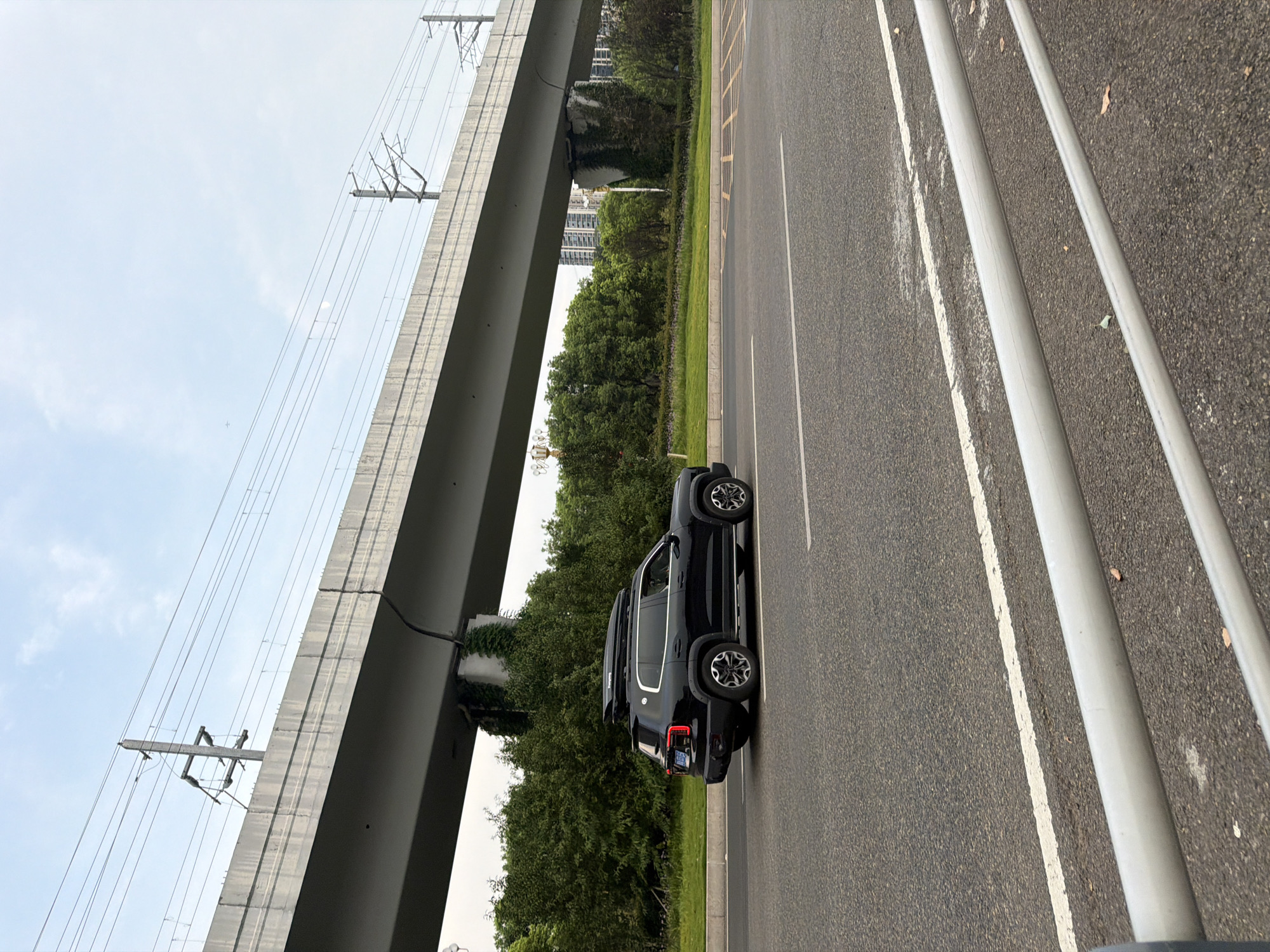 A black SUV with a roof rack is driving on a road in Jiangjin, China. The vehicle is in the middle lane of a multi-lane road, and it is moving from left to right in the frame. The road is asphalt and has white lane markings. In the background, there is a large elevated concrete structure, which appears to be an overpass or a viaduct, with overhead power lines. There are trees and bushes on the side of the road, and in the distance, there are some buildings. The sky is overcast. The overall scene depicts an urban or suburban environment with transportation infrastructure.