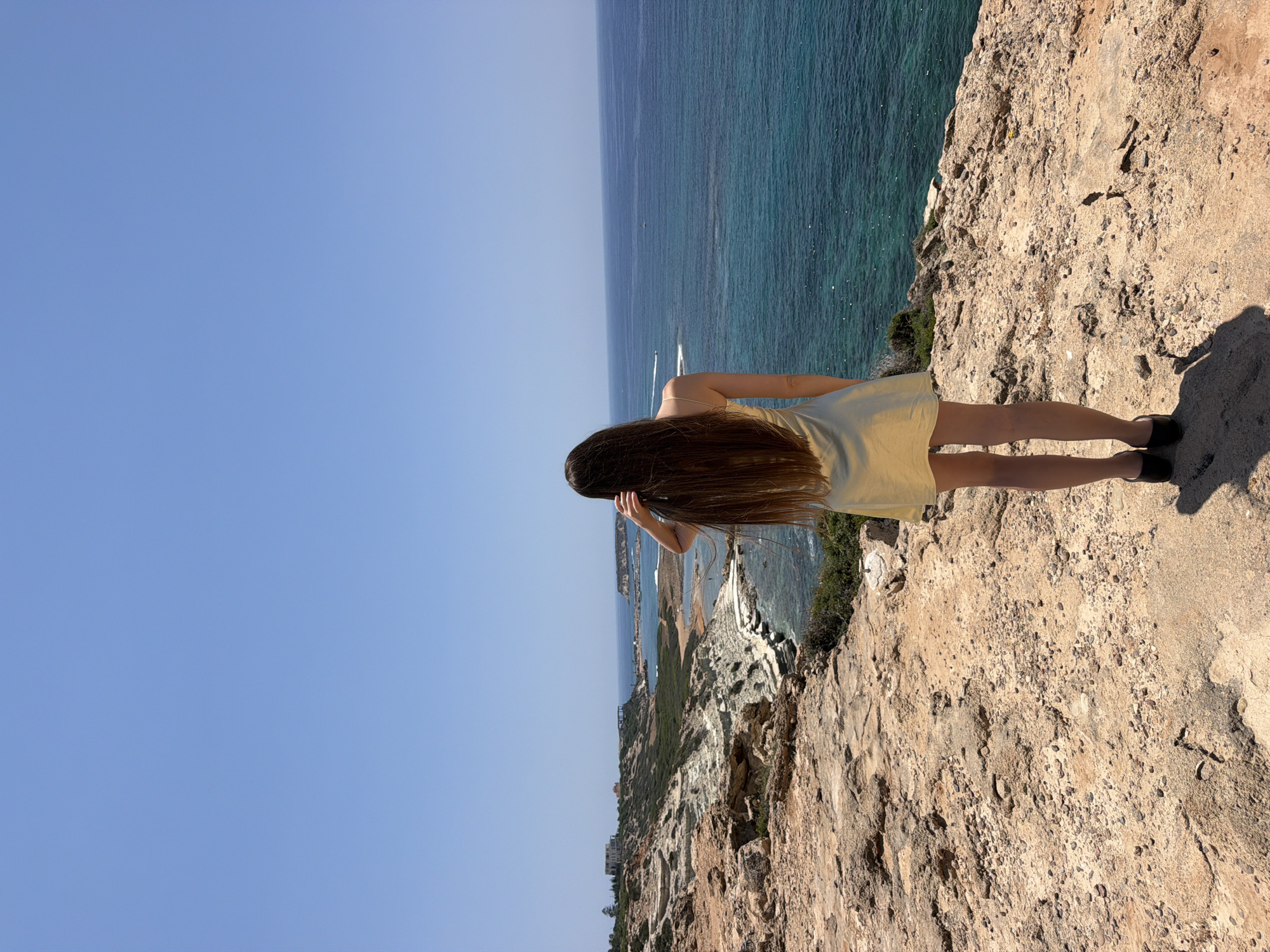 A person stands on a cliff overlooking a body of water. The person has long, dark hair and is wearing a yellow dress. The sky is a clear blue, suggesting it is a sunny day. The water is a vibrant turquoise, with a gentle ripple suggesting calm conditions. The cliff face is rocky and arid, with sparse vegetation in some areas. In the distance, there is a coastline with buildings and more vegetation. The image appears to have been taken during daylight hours. The person is looking out towards the horizon, creating a sense of contemplation or awe at the natural scenery.