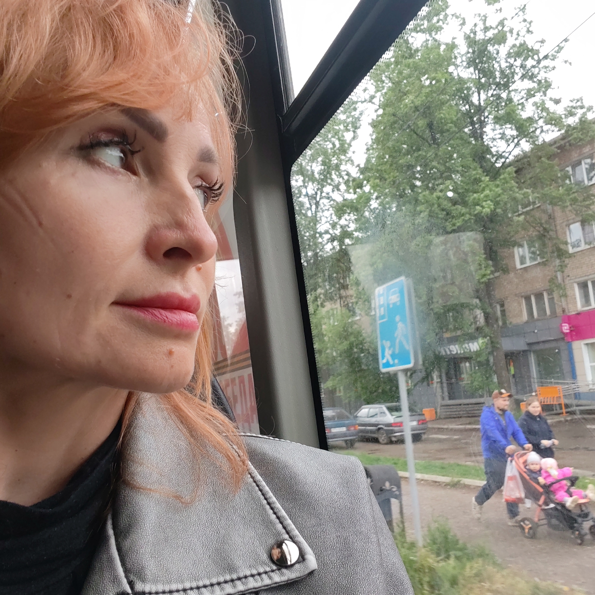 A woman with reddish hair and wearing a gray leather jacket is looking out the window of a vehicle, likely a bus, as it travels along a street in Chaikovsky, Russia. Outside the window, a pedestrian crossing sign depicting a bus and people walking is visible. Further in the background, a brick building with windows and signage can be seen. On the sidewalk, a man wearing a blue jacket is pushing a stroller with two children, while a woman in a dark jacket walks alongside them. Cars are parked nearby. The overall environment suggests an urban setting, possibly a residential area with commercial establishments. The weather appears overcast.