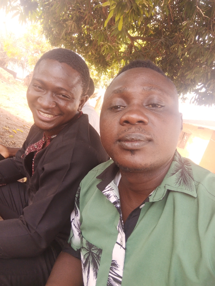 Two dark-skinned men pose for a selfie outdoors under trees. The man on the left smiles broadly, wearing a black shirt with red and white embroidery around the collar. The man on the right, holding the camera, wears a green shirt with a black and white tropical print. Both men have short, dark hair. The background features trees and a glimpse of a building. The lighting is bright, with some areas slightly overexposed.