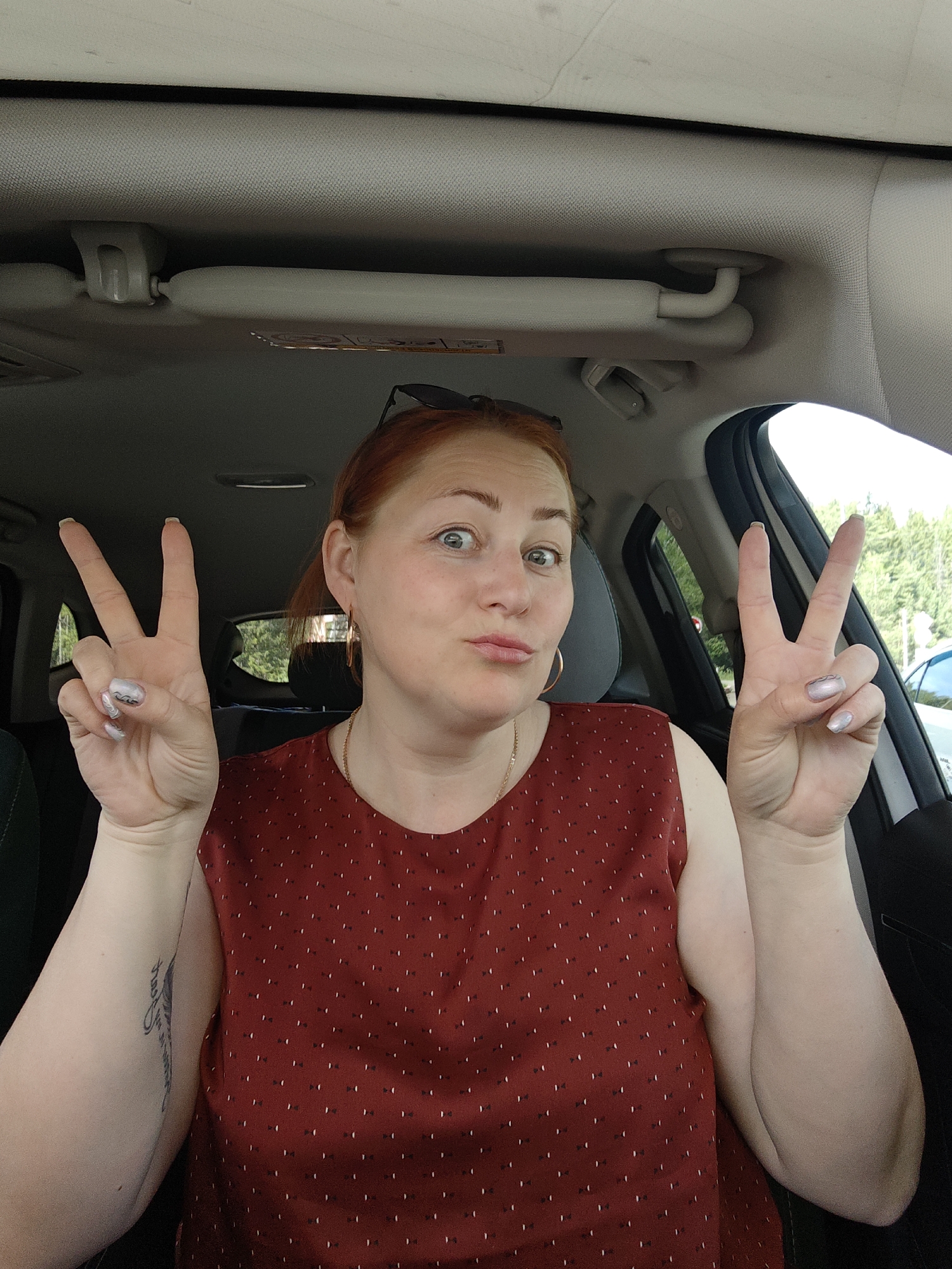 A light-skinned woman with red hair sits in the driver's seat of a car, holding up both hands with her fingers shaped into a "peace" or "victory" sign. She wears a maroon, sleeveless top with small, white polka dots. A gold necklace and gold hoop earrings are visible. Her nails are painted a pearlescent white. The car's interior is visible, including the sun visor, ceiling, and part of the dashboard. Through the open driver's side window, trees and a glimpse of another car are seen. The car is possibly located in or near Chaikovsky, Russia.