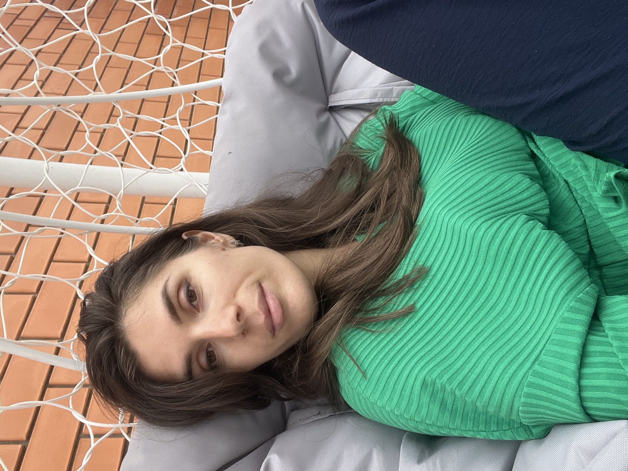 A young woman with brown hair is reclining on a gray pillow inside a hammock-style chair. She is wearing a green, ribbed shirt and a dark blue garment is visible above her. The hammock's white frame and netting are visible, as is a brick-colored ground beneath it. The location is outdoors, potentially in Kaspiysk, Russia.